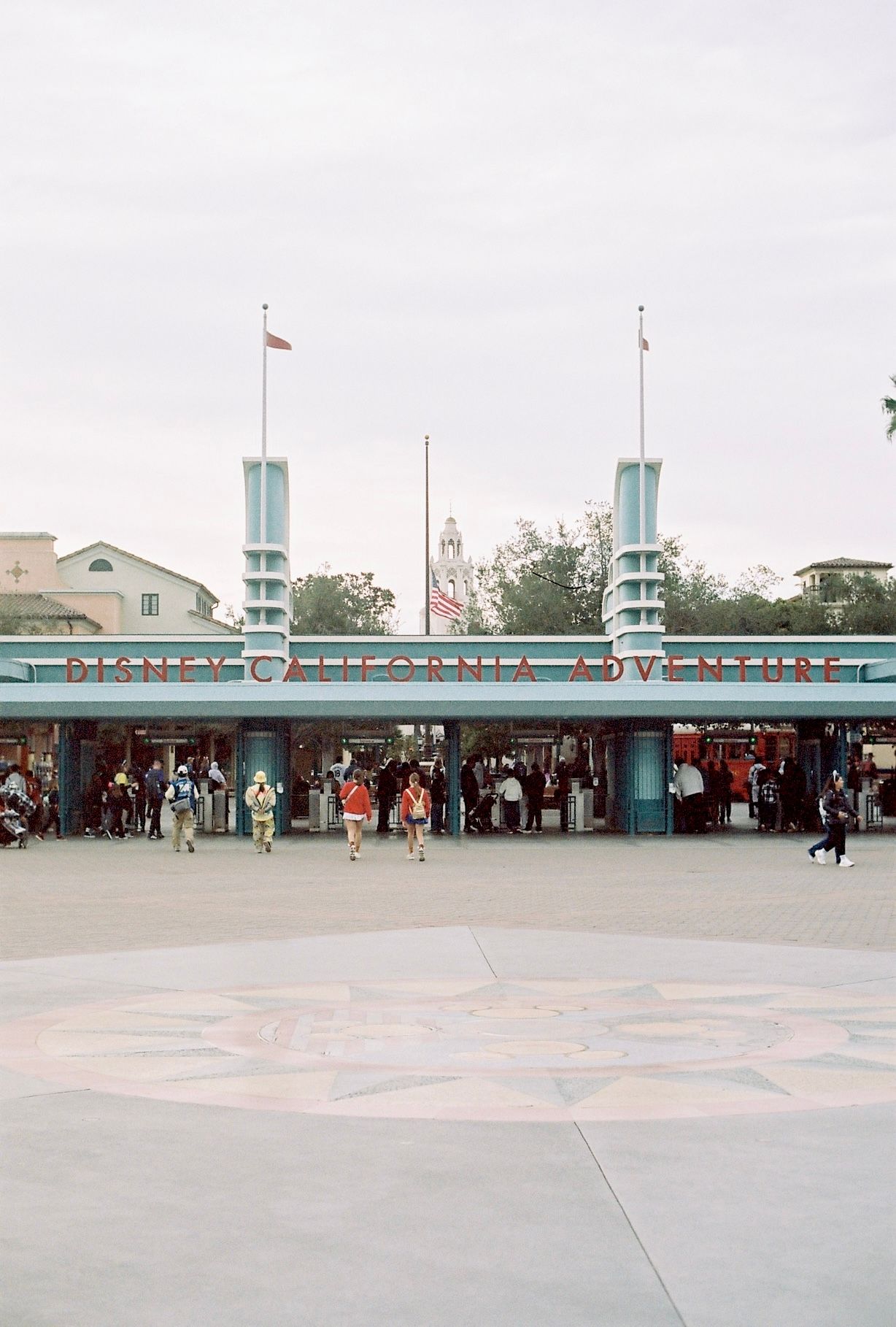 A long horizontal view of a retro-style theme park entrance with turquoise architectural elements and crowds gathering.