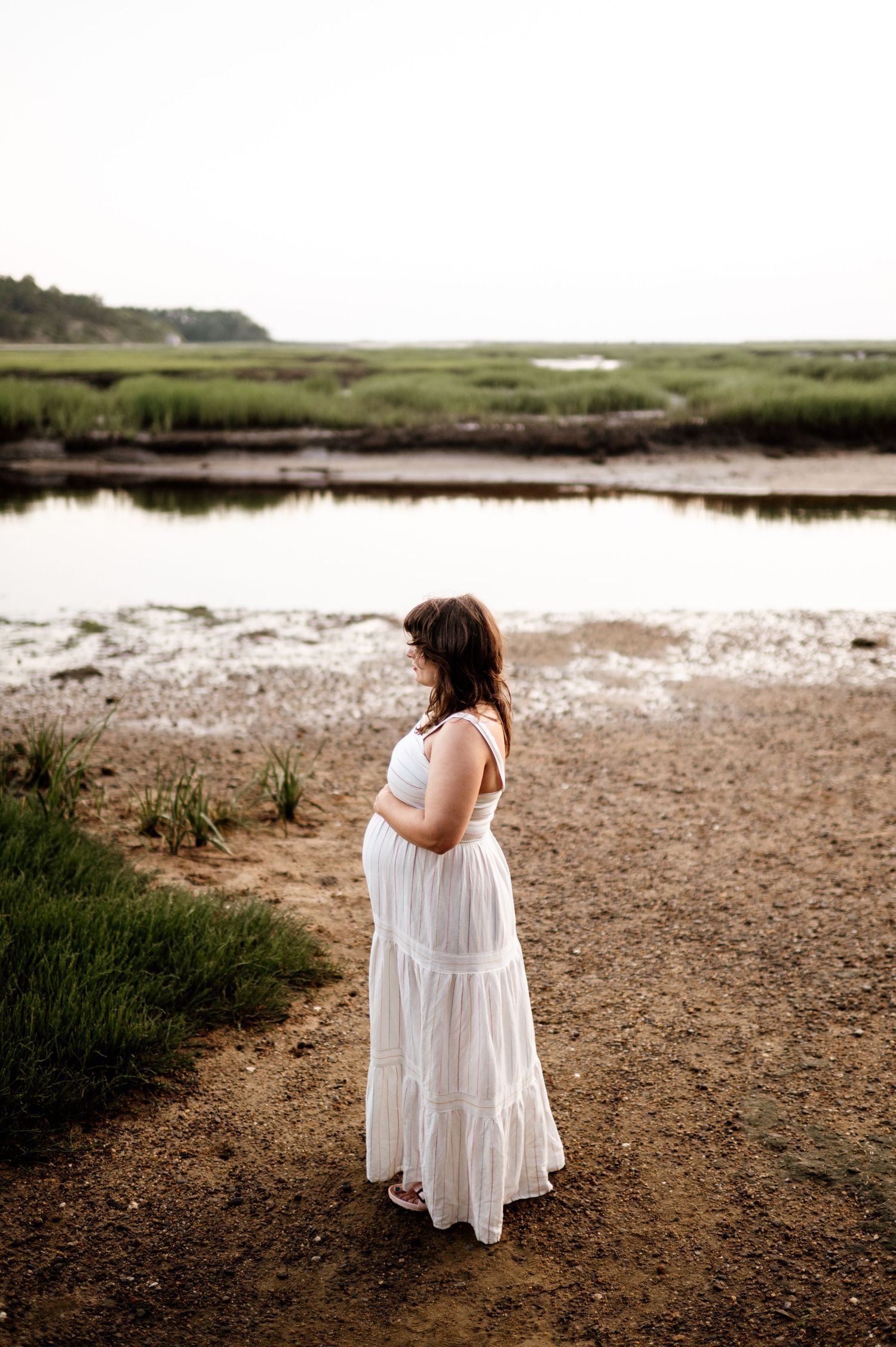 Person in flowing white dress walking along marshy coastline at dusk.