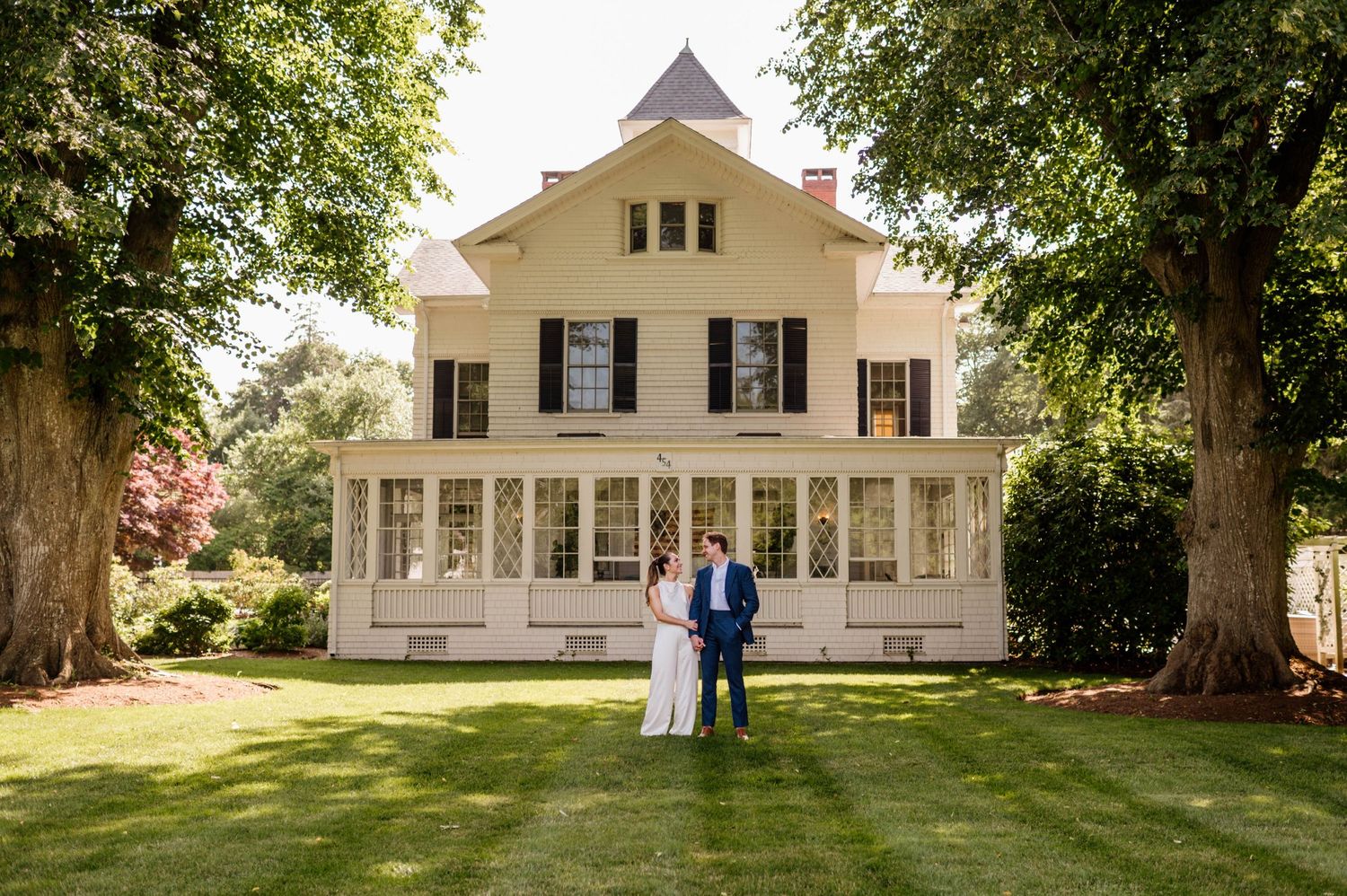 A row of white farmhouse-style buildings with wrap-around porches set against lush green trees on a sunny day.