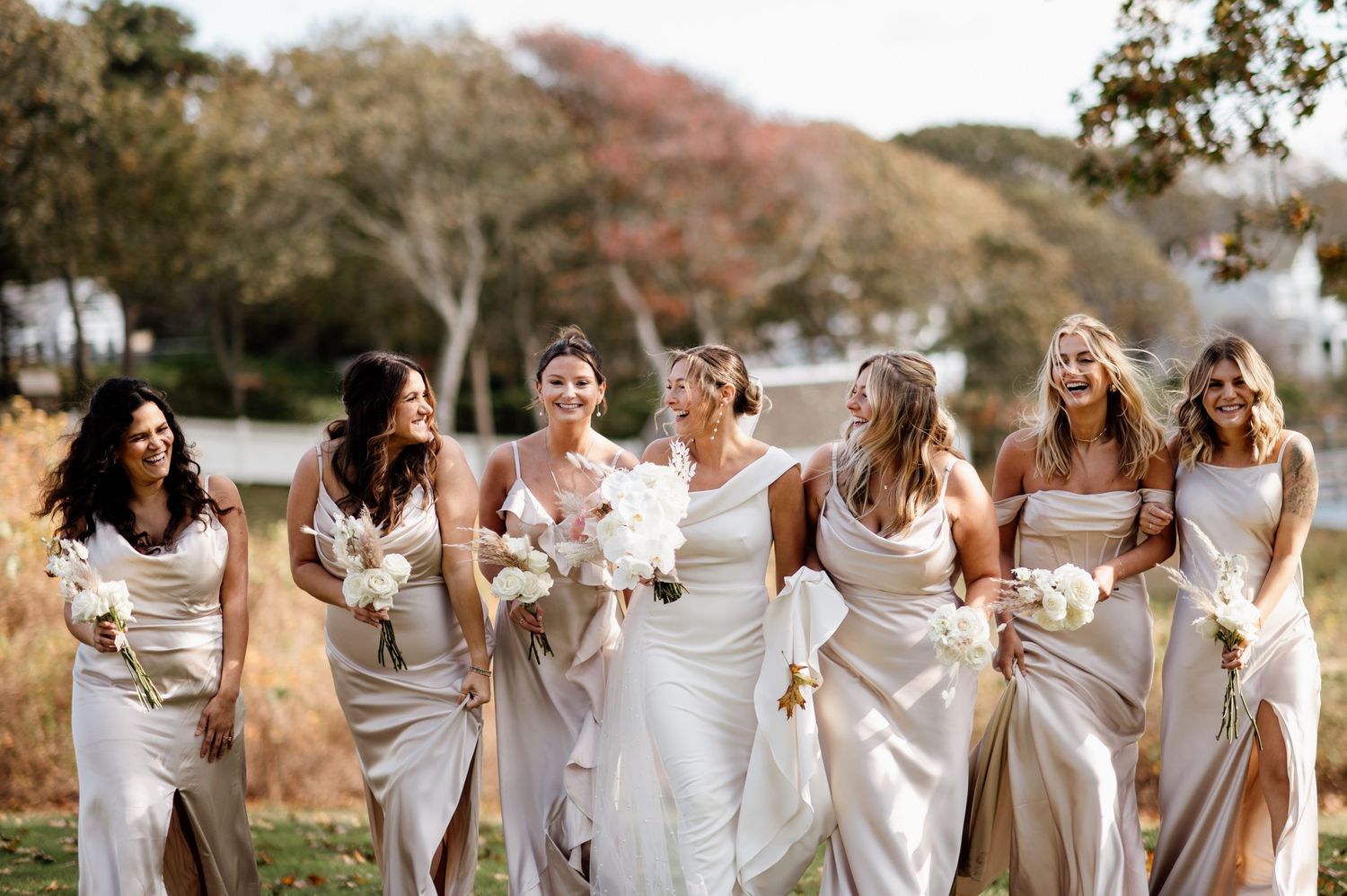 Group of bridesmaids in white dresses laughing together outdoors against a natural autumn backdrop.