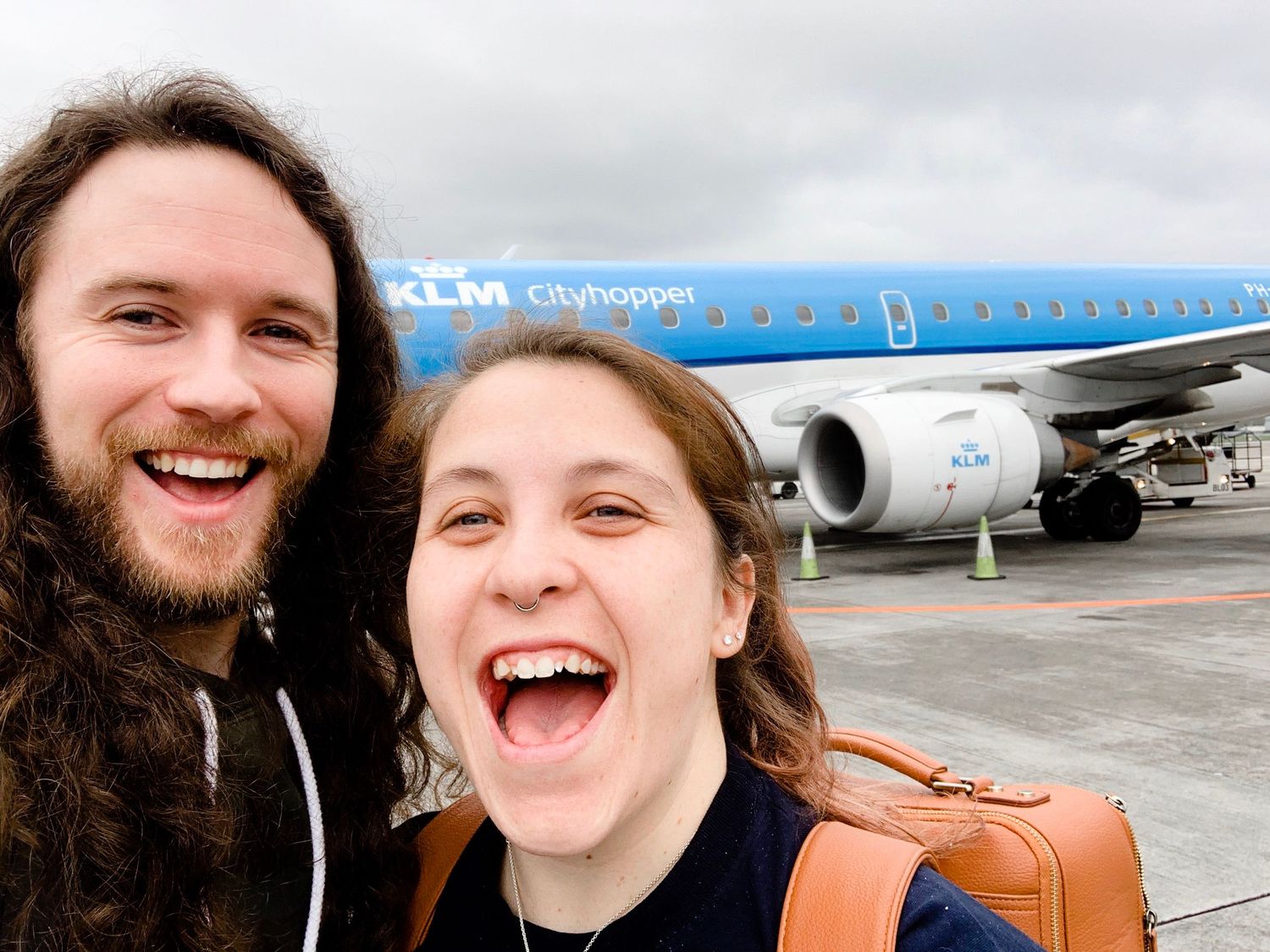 Two excited travelers take a selfie in front of a blue KLM airplane on the tarmac.