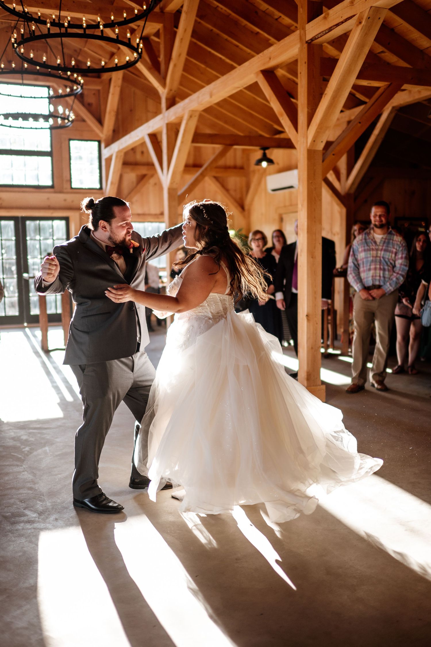 Wedding couple dances in sunlit rustic barn venue with exposed wooden beams and string lights.
