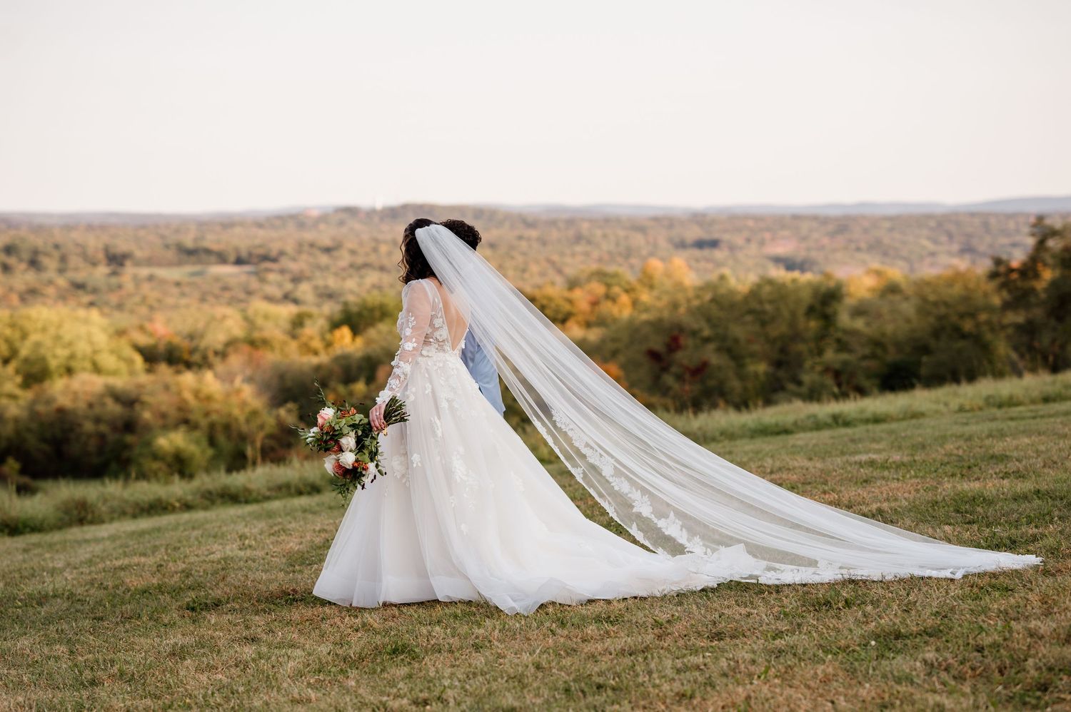 A long flowing white wedding dress and veil captured against a scenic autumn hillside landscape at sunset.