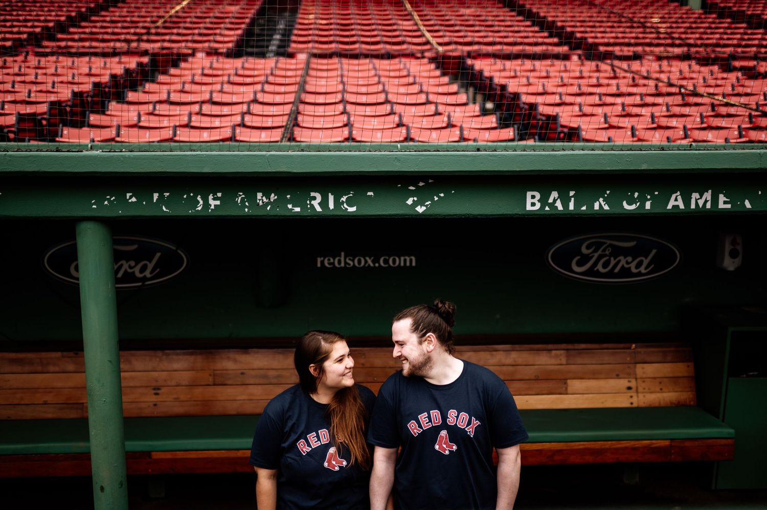 A couple shares intimate moments in front of empty red stadium seats at a baseball field.