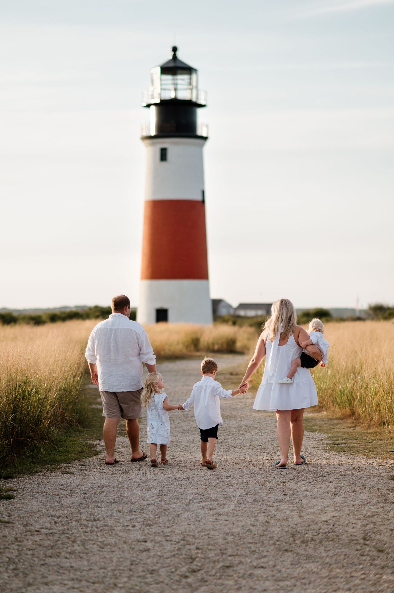 Family walking toward lighthouse on sandy path with tall grass and warm evening light.