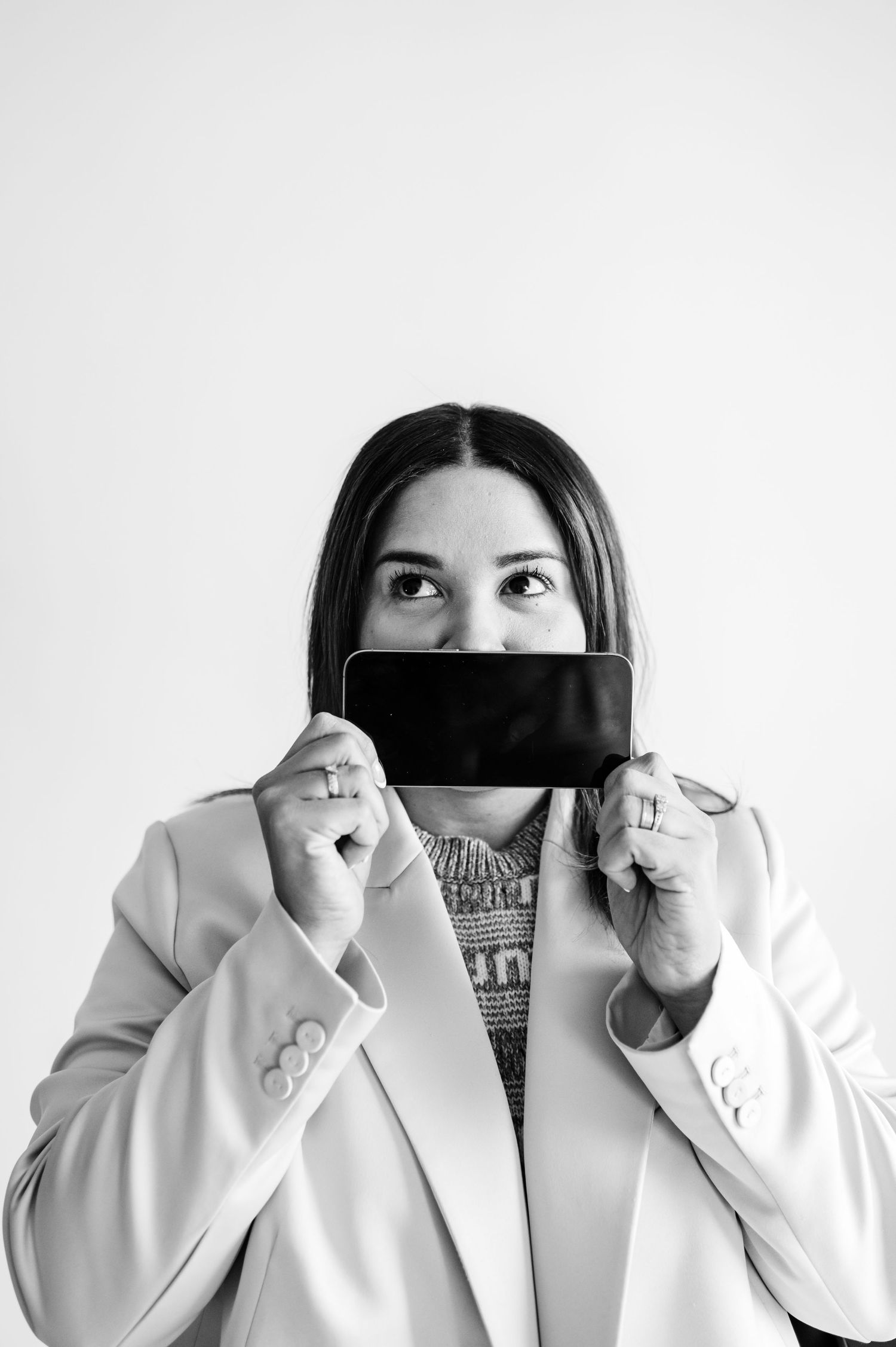 A person in a white blazer holds a black strip of fabric across their face in a dramatic black and white portrait.