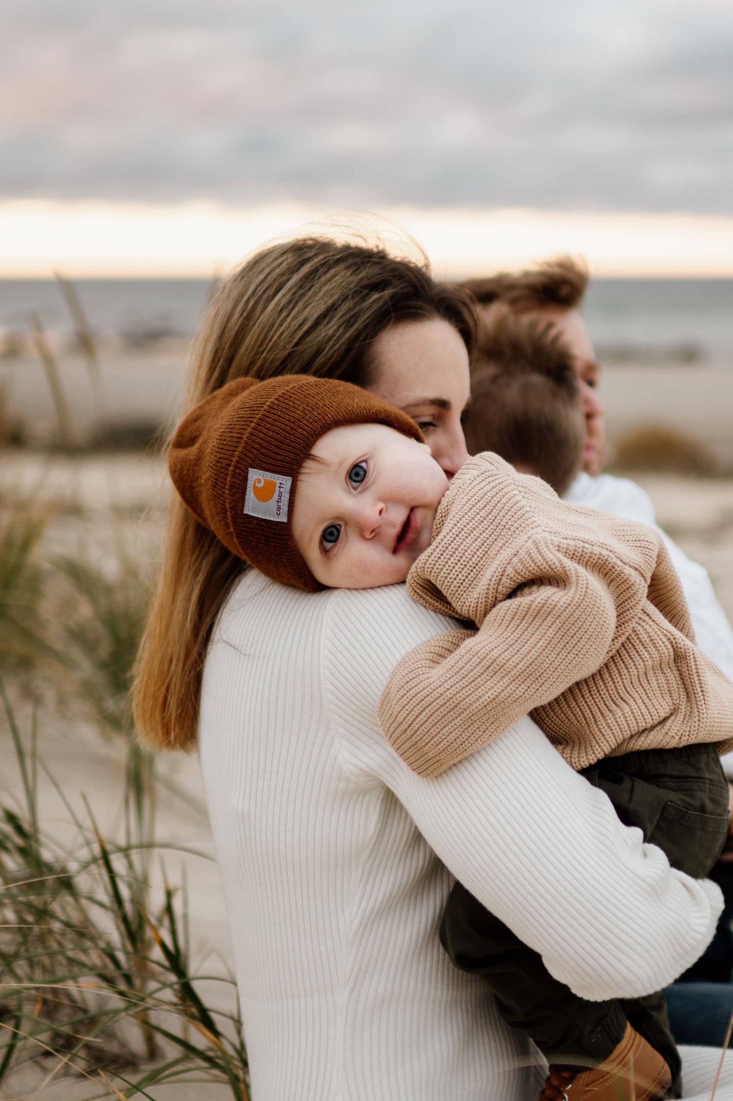 A couple in cozy sweaters embraces while sitting together on a beach during sunset.