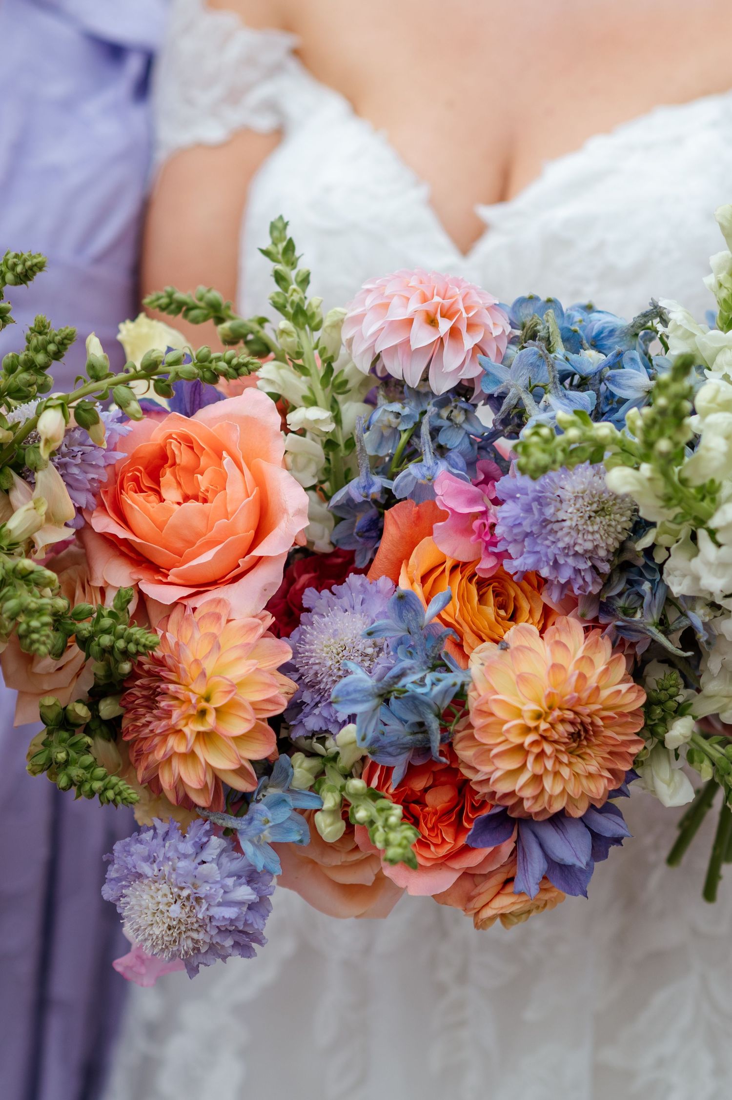 Bride holding a vibrant wedding bouquet with peach dahlias, blue hydrangeas, coral roses, and purple scabiosa flowers.