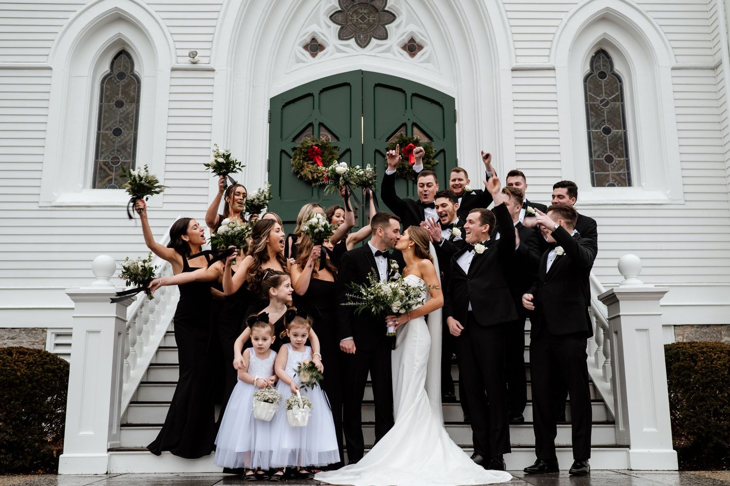 Wedding party celebrates joyfully outside a white church with Gothic arched doorways.