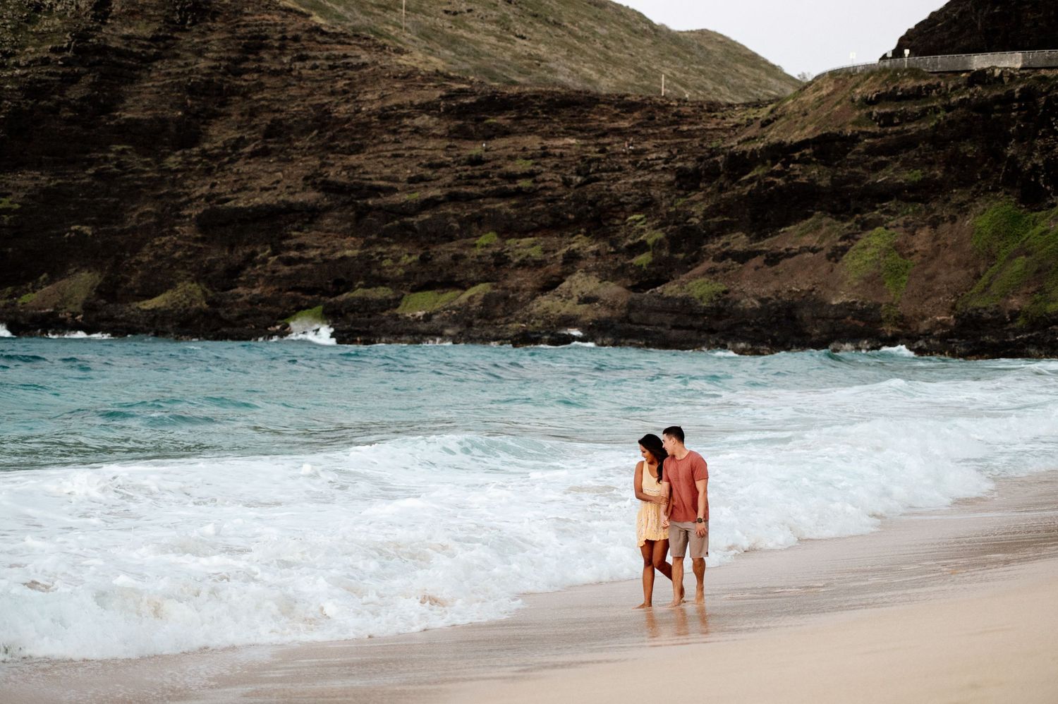 A couple walking along a tropical beach with dramatic cliffs and turquoise waters.