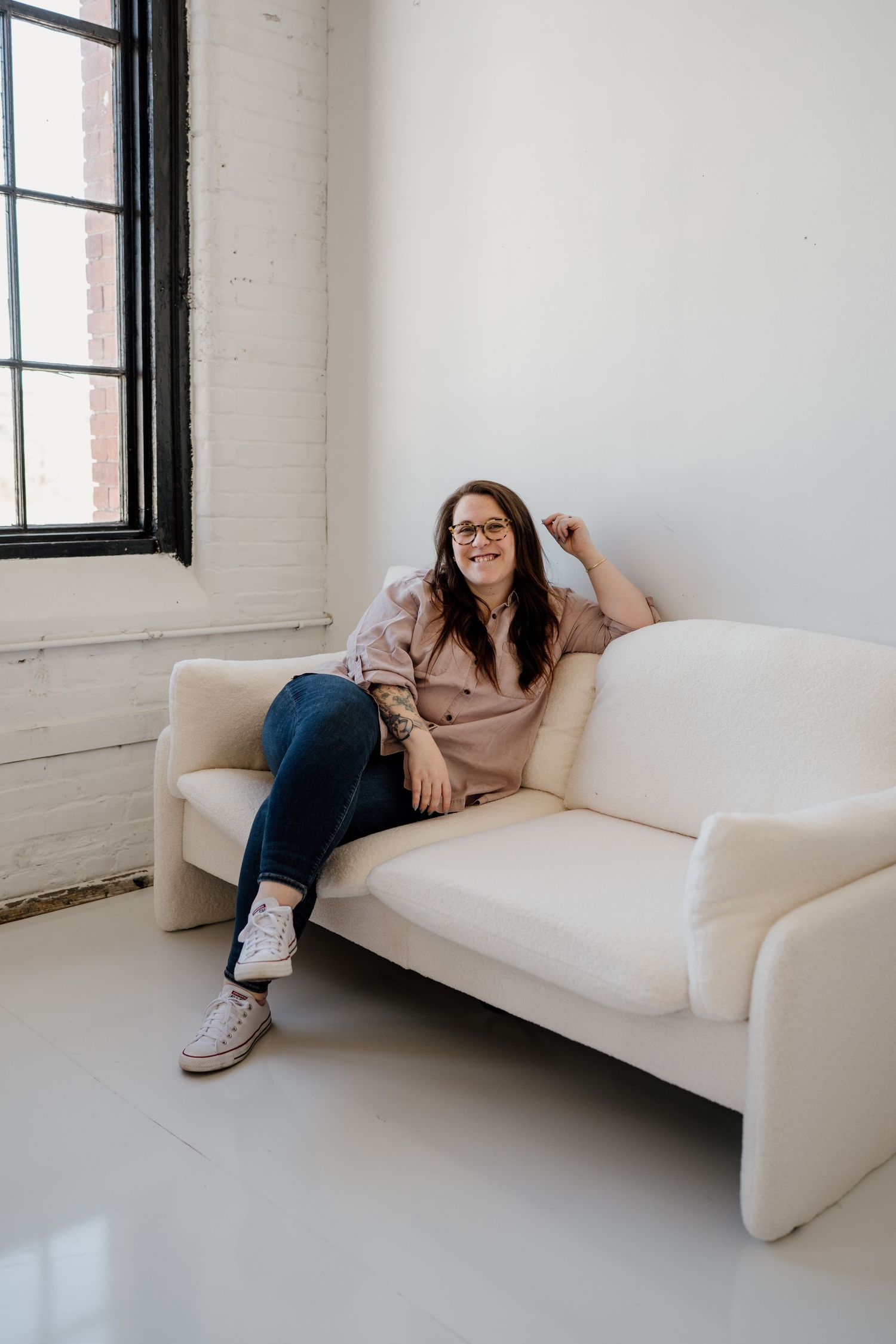 A person wearing glasses and jeans reclines on a white modern sofa in a bright minimalist room with large windows.