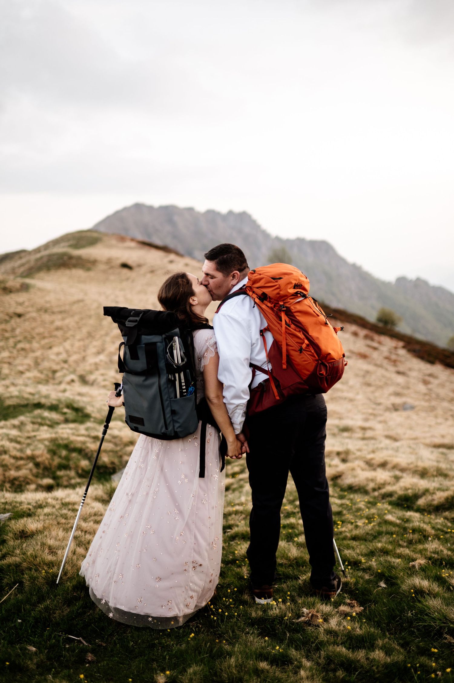 A couple in wedding attire share an intimate moment while hiking in the mountains with backpacks.