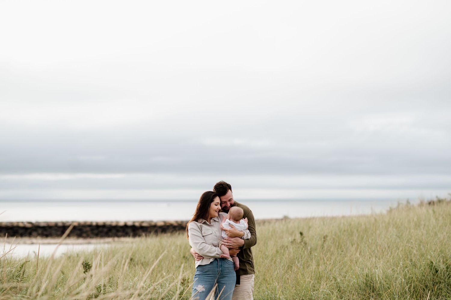 Couple embracing in a grassy field overlooking the ocean on an overcast day.