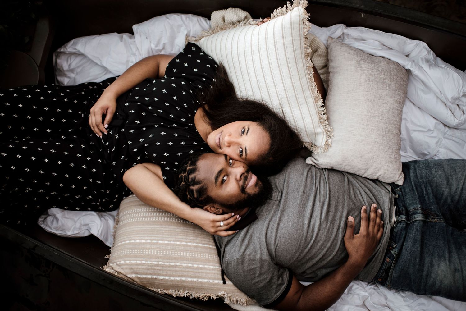 A couple lying on white bedding among decorative pillows in an intimate lifestyle portrait.