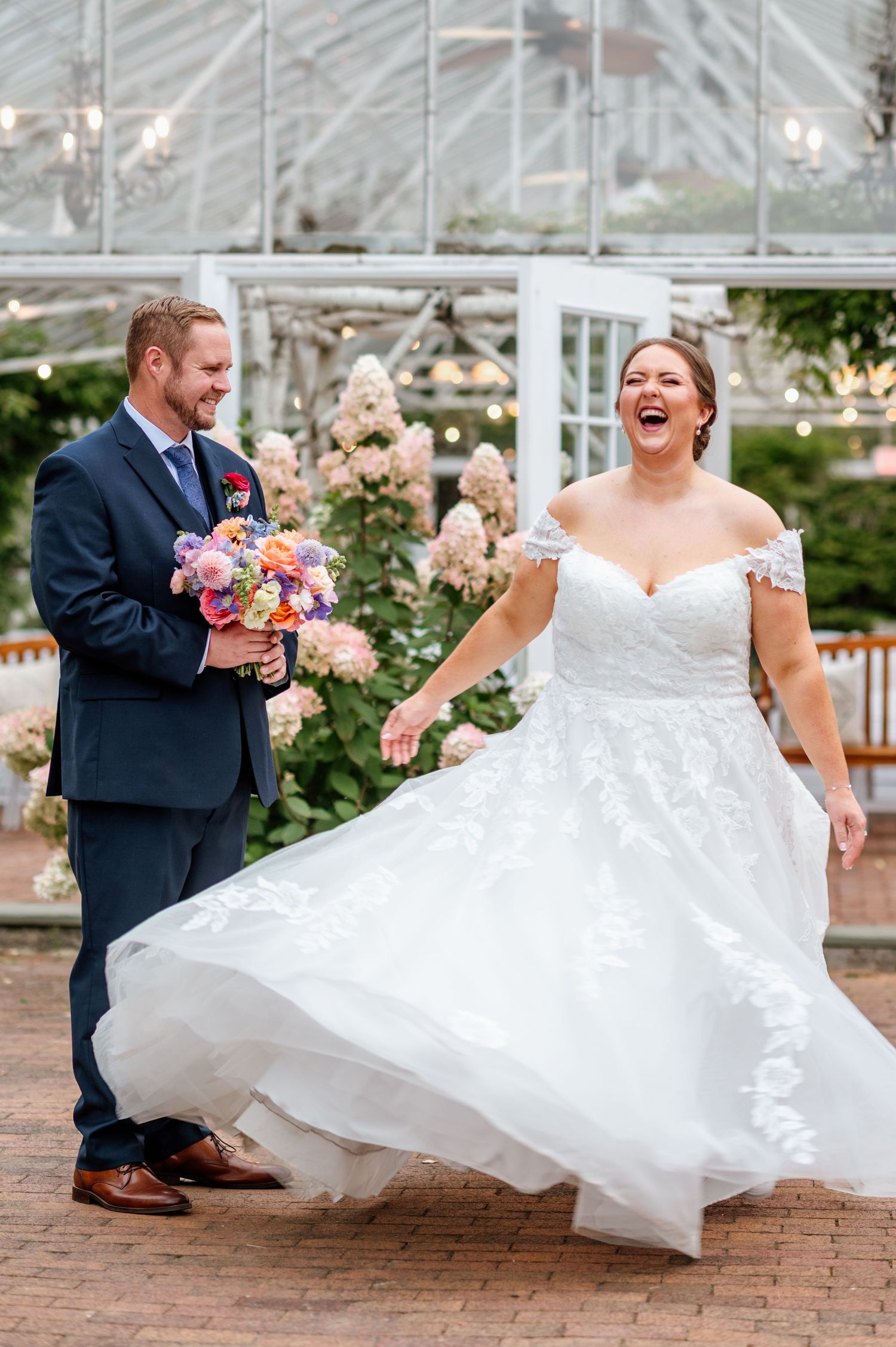 Joyful bride in lace off-shoulder gown twirling with groom holding colorful bouquet at elegant greenhouse wedding.
