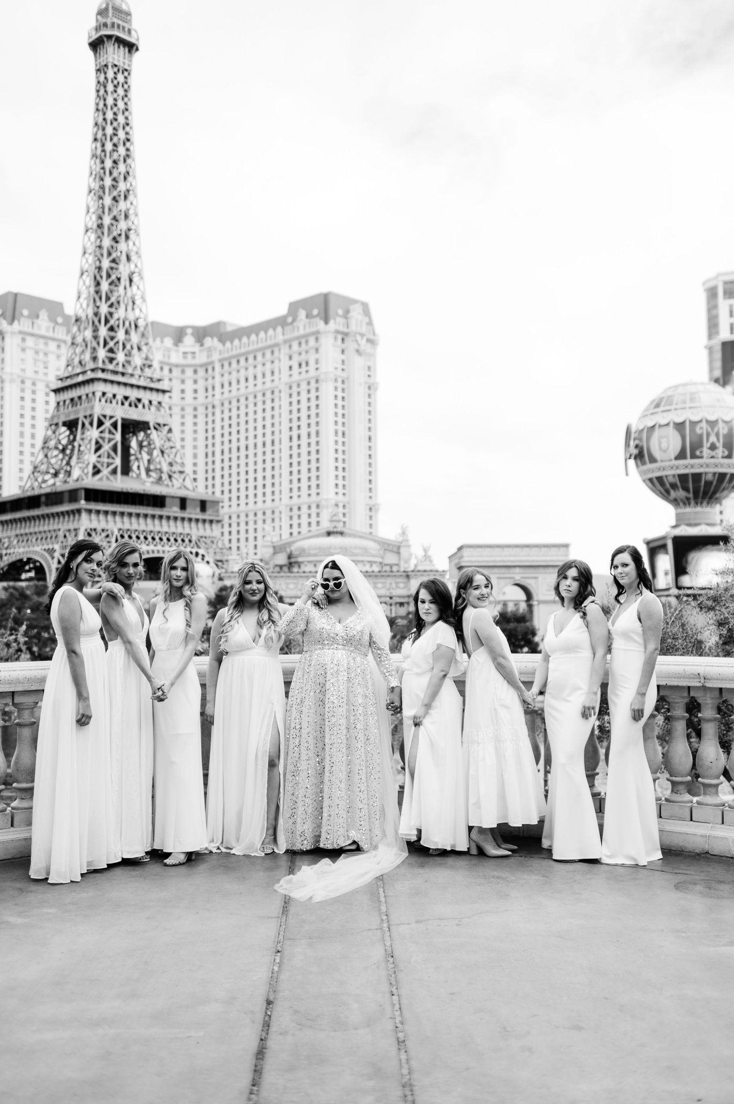 A bridal party in white dresses poses together on a balcony with the Paris Las Vegas hotel and Eiffel Tower replica in view.