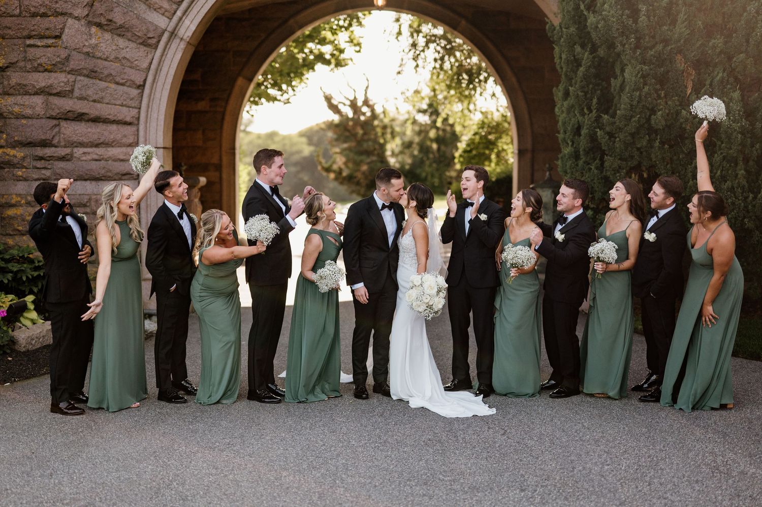 Wedding party in elegant green dresses and black tuxedos poses together under stone archway at sunset.