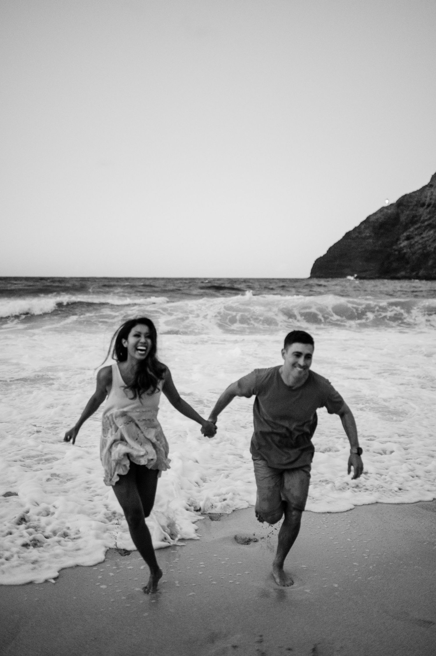 Playful couple runs from ocean waves on beach during black and white photoshoot.