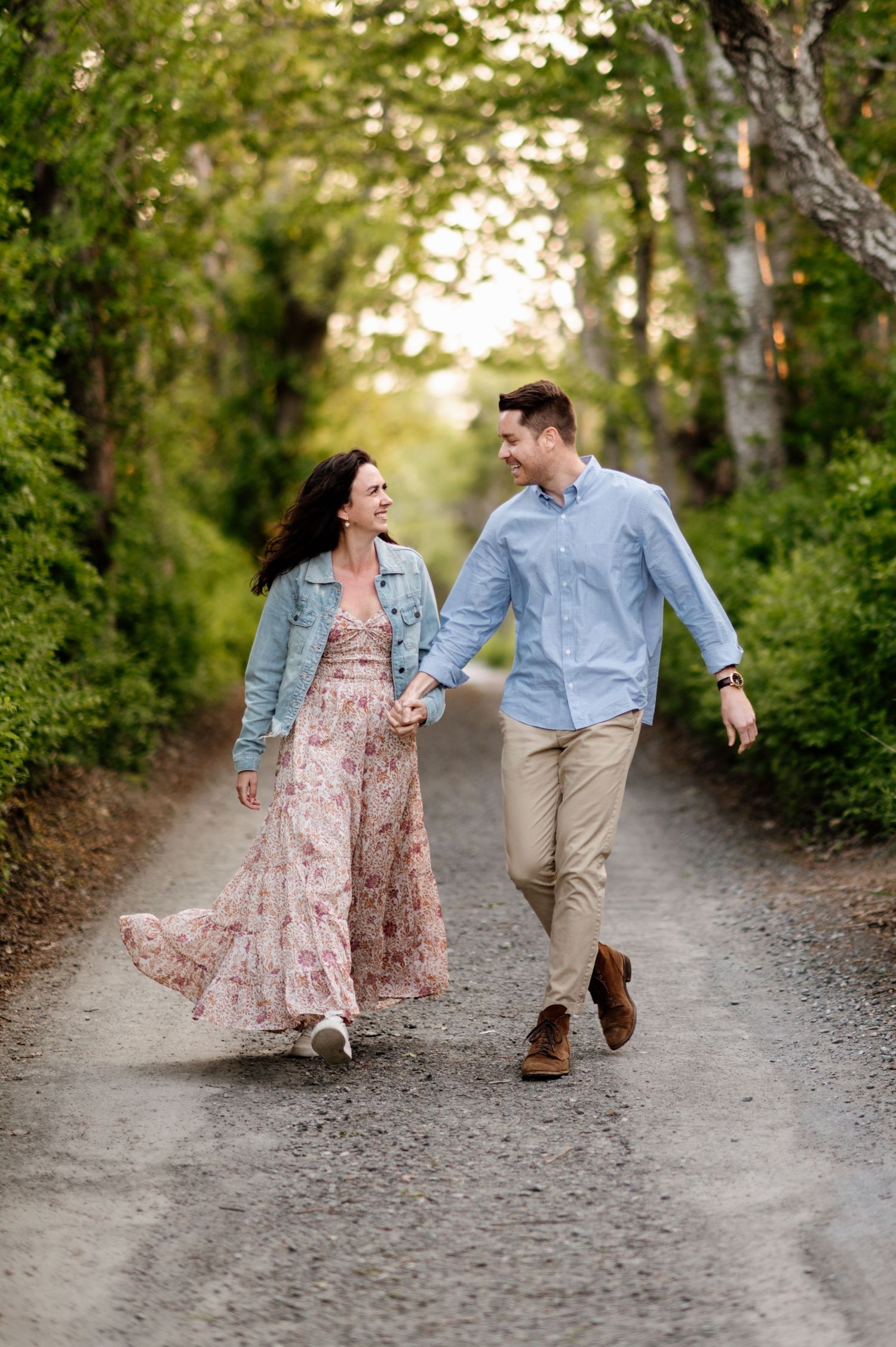 Romantic couple walking and dancing together on a tree-lined path in casual attire during golden hour.