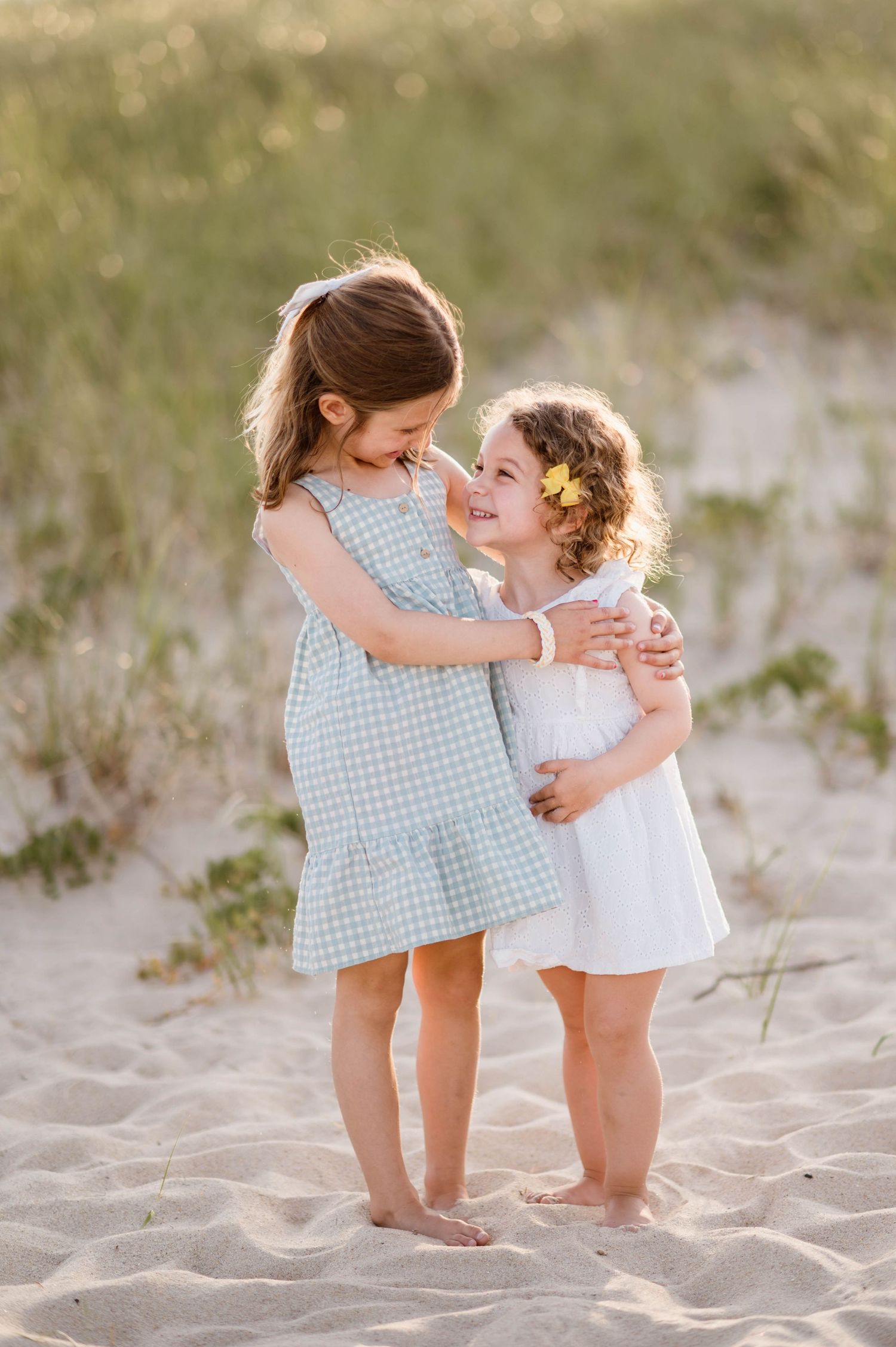 Two small children in summer outfits hugging on a sandy beach.