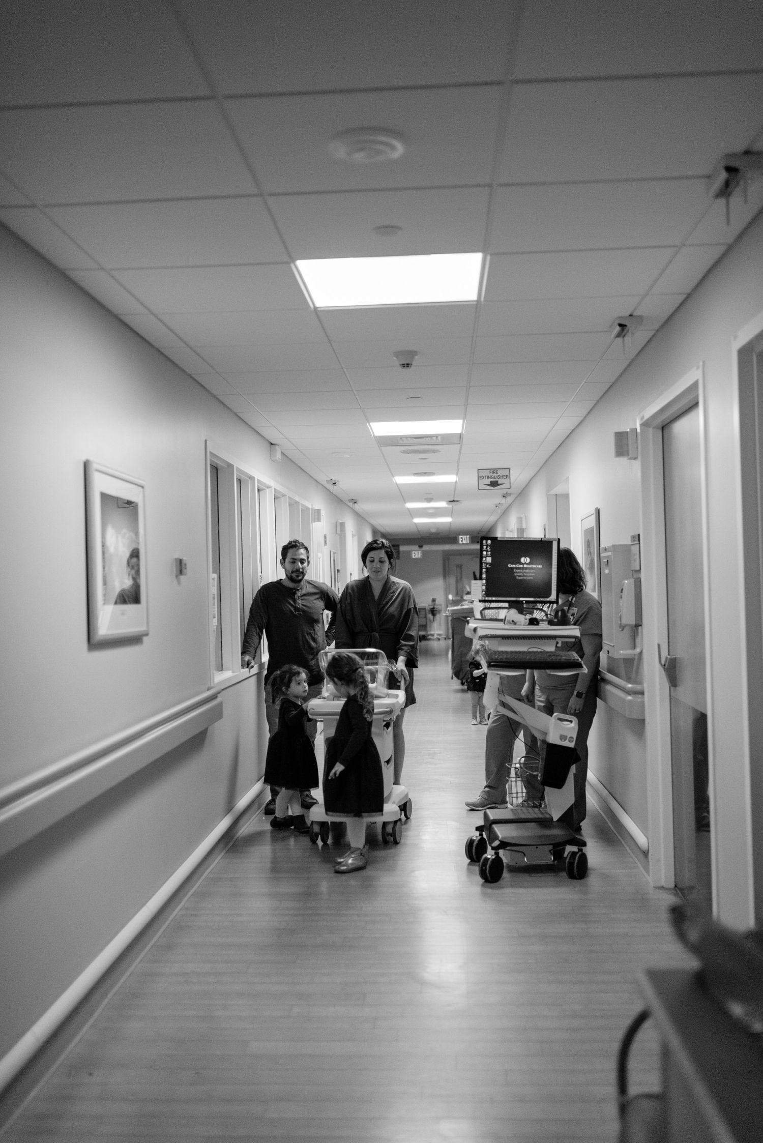 Black and white hospital hallway with people walking and medical equipment.