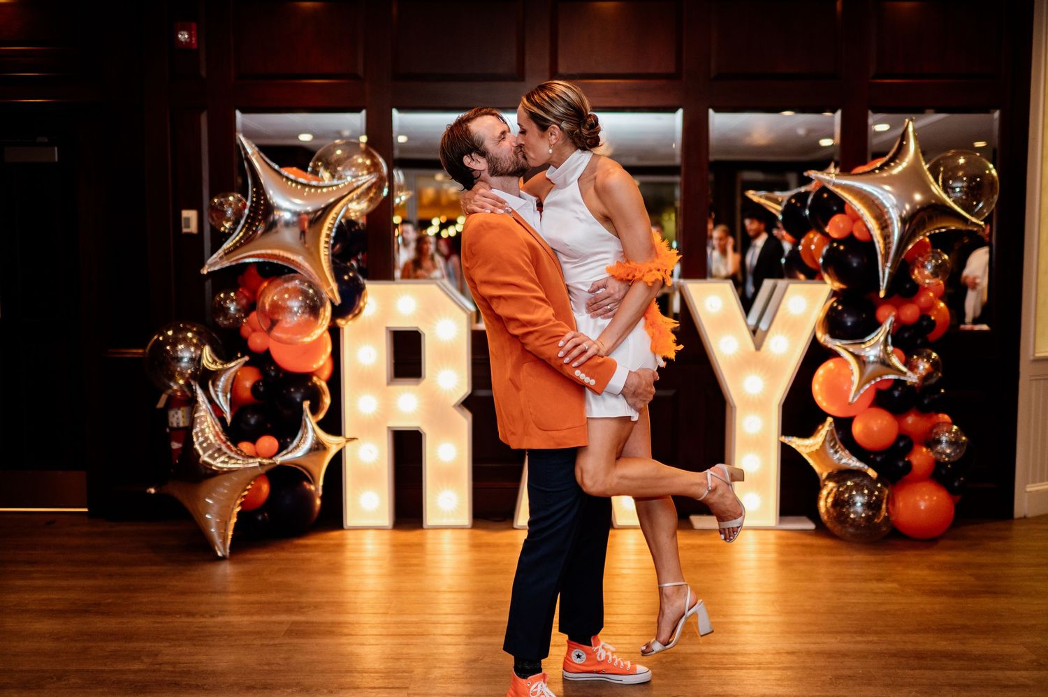 A couple embraces in front of illuminated JOY letters with gold star balloons and festive decorations.