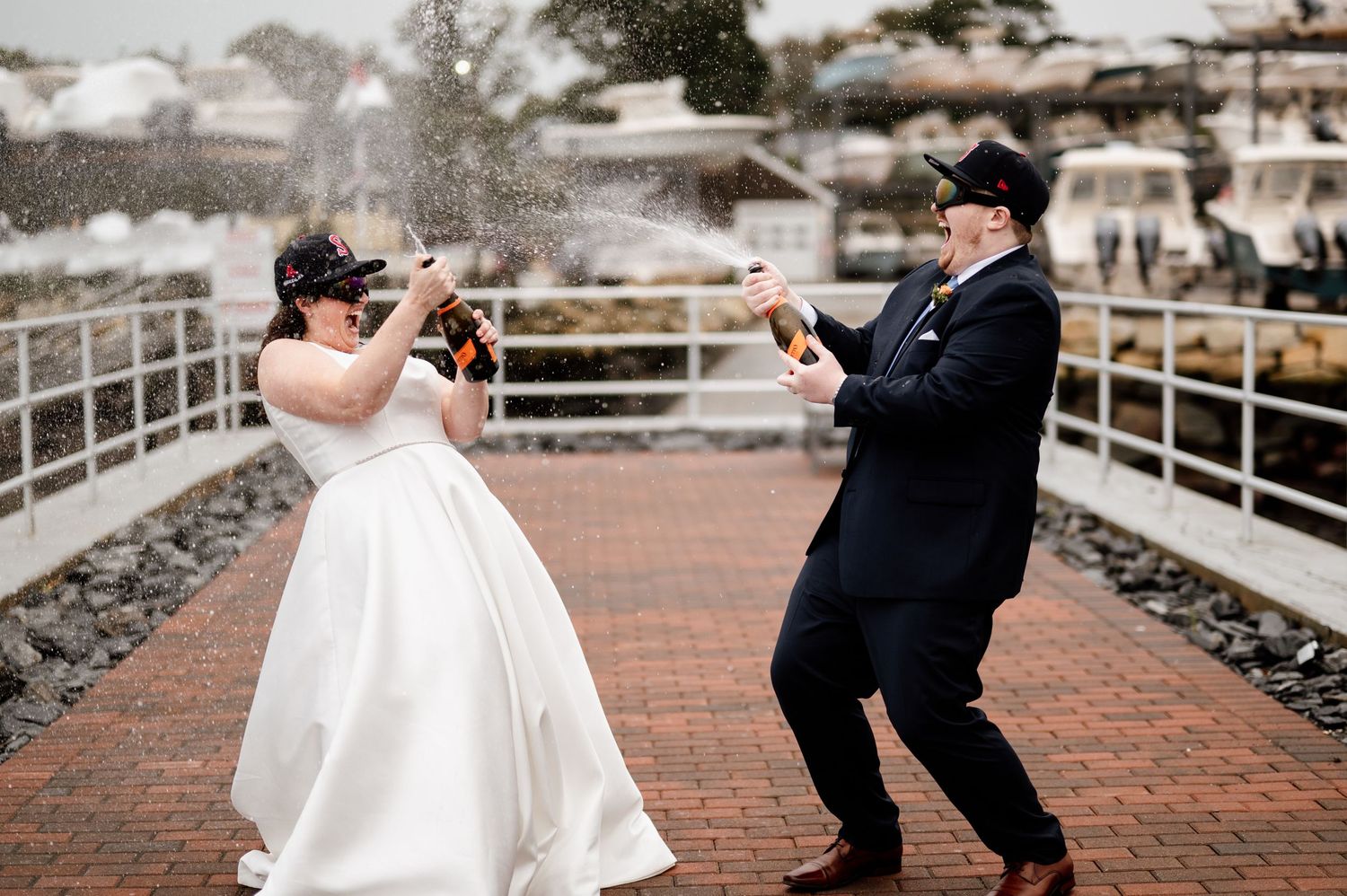 A couple in wedding attire dances and twirls on a brick walkway near a marina.
