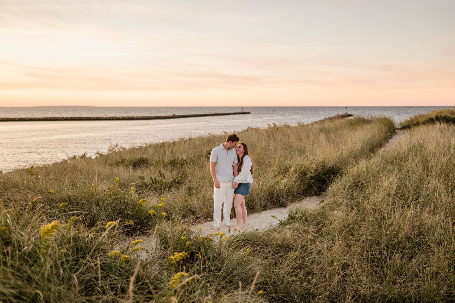 Couple walking along scenic coastal path through tall grass at sunset by the ocean.