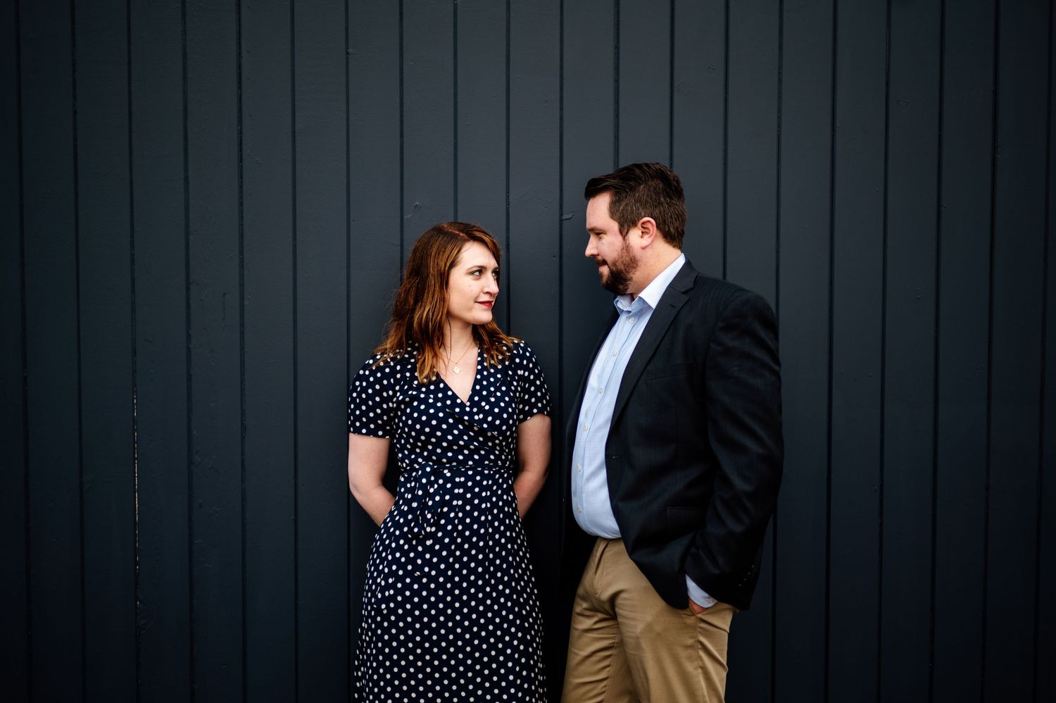 Romantic engagement session with couple in polka dot dress and blazer against dark wooden wall.