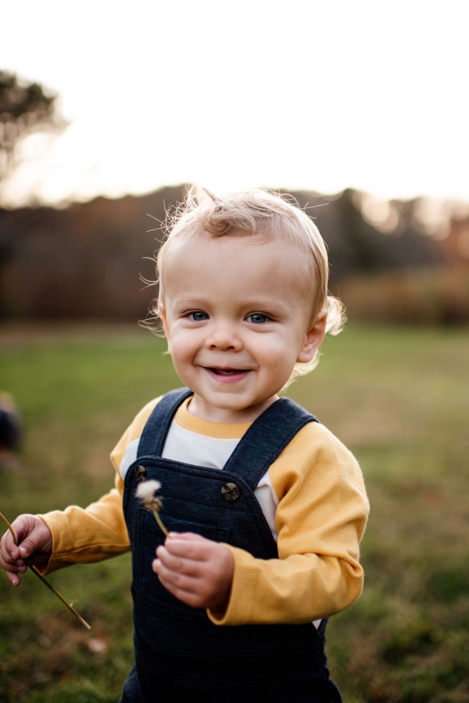 A toddler in overalls and yellow shirt smiles while playing outdoors at sunset.