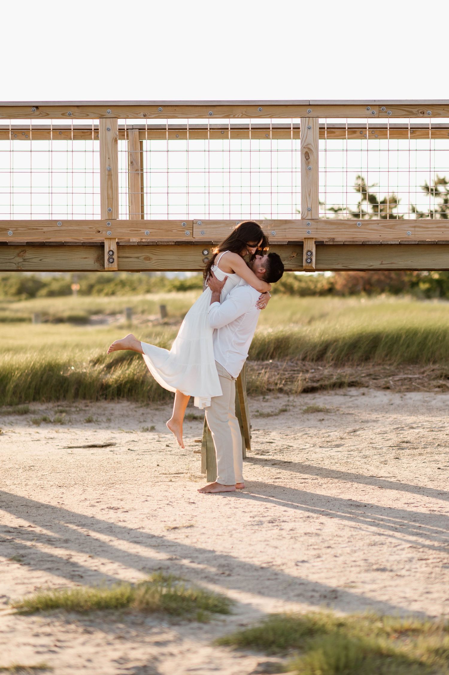A couple in white clothing performs a romantic dance lift pose on a sandy beach at sunset.