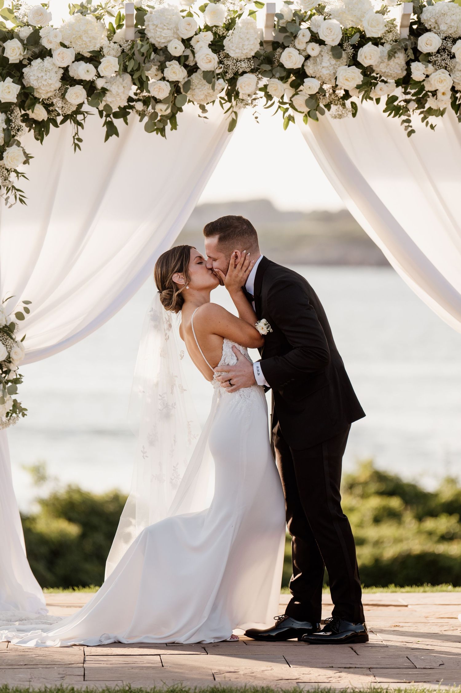 A romantic moment under a white floral arch during an outdoor wedding ceremony.