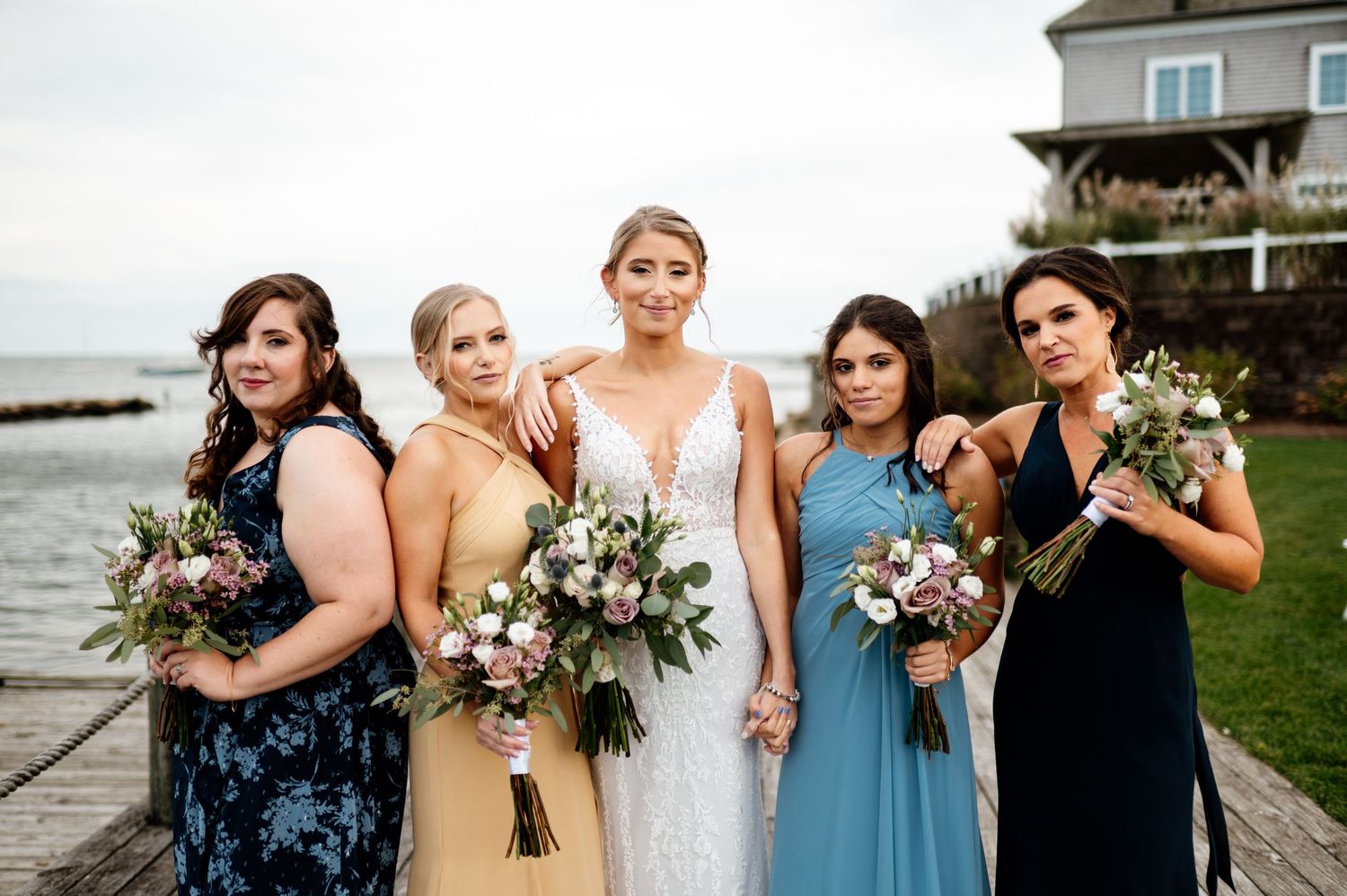 Bridesmaids in blue dresses holding floral bouquets pose together outdoors.