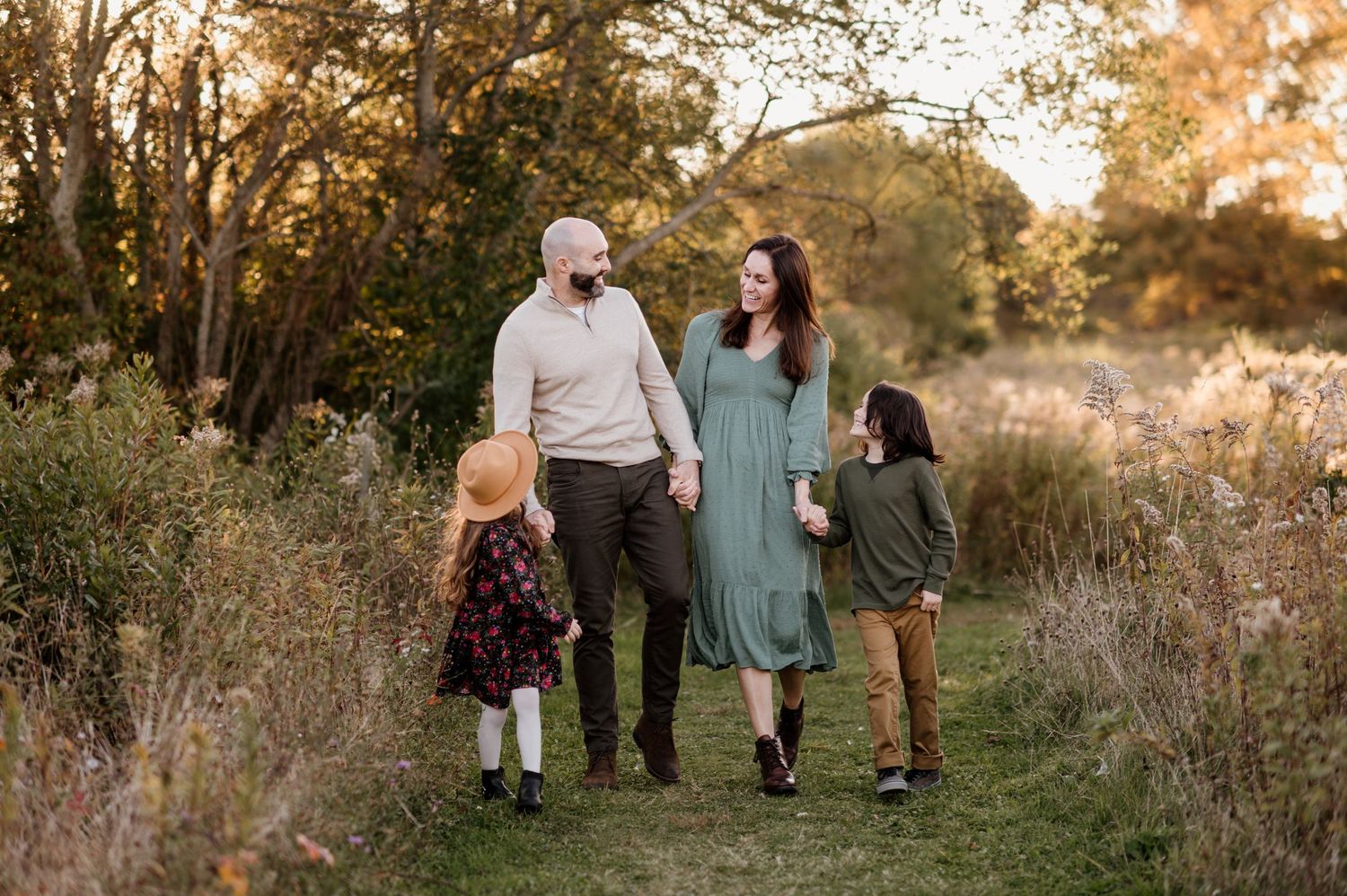 Family of four walking together on a scenic autumn path surrounded by golden foliage and warm sunlight.