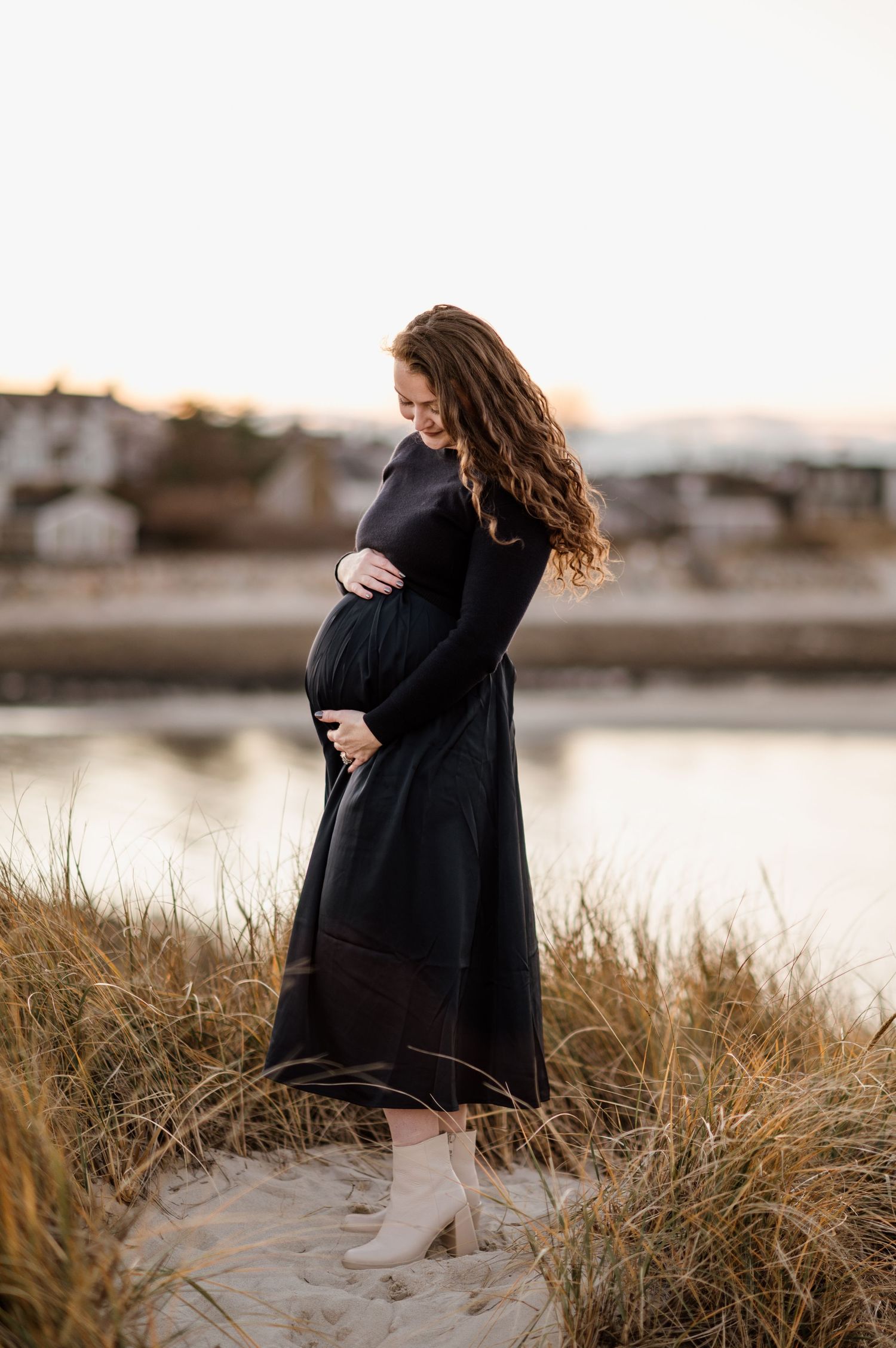 Silhouette of pregnant woman in black dress standing by water during golden hour.