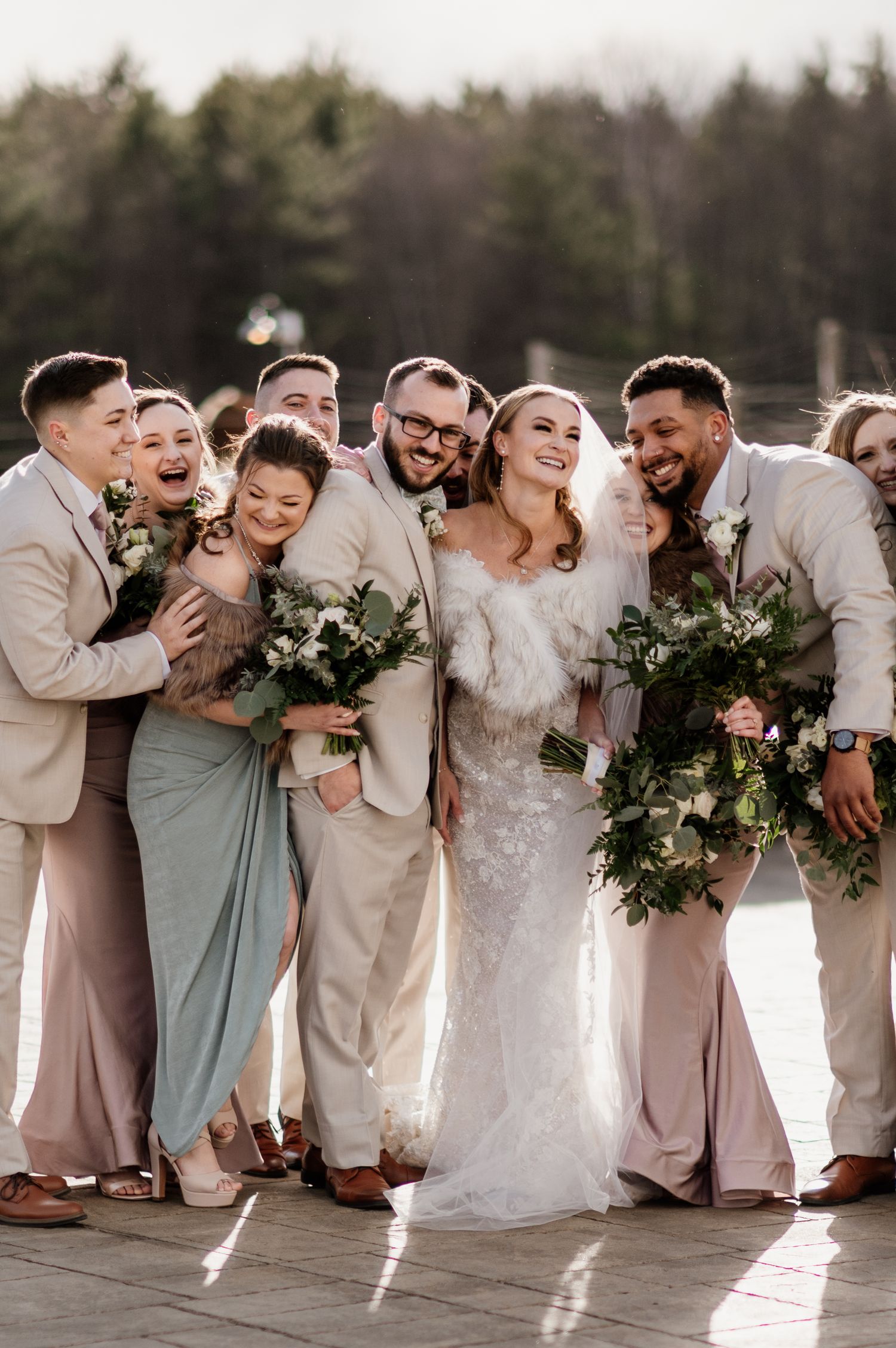 Wedding party in light beige suits and sage dresses laughs together in a candid group photo.