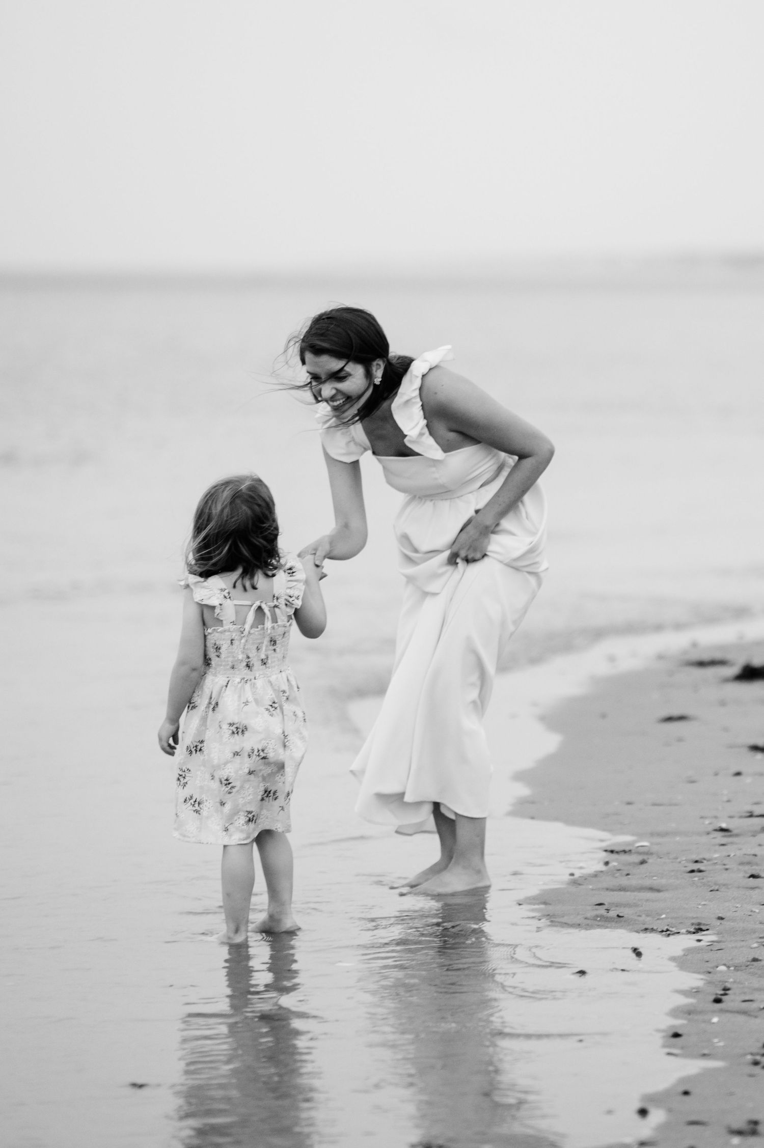 Black and white photo of a parent and child walking together along the shoreline at the beach.