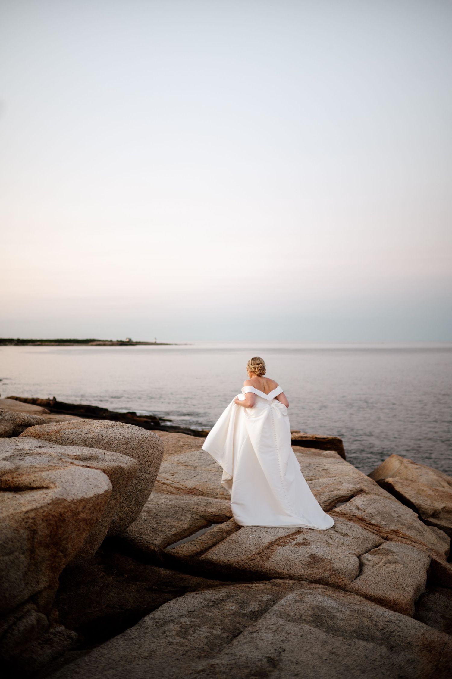A white wedding dress flows gracefully against a serene coastal landscape with large rocks and calm waters at sunset.