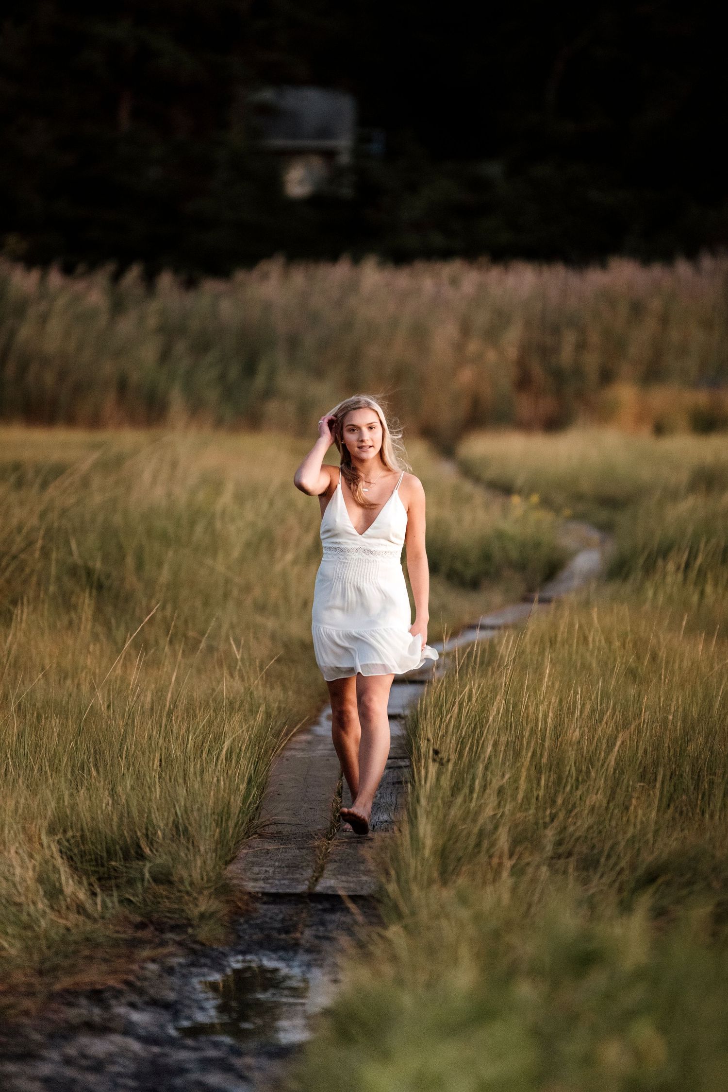 Person in white dress sits thoughtfully on wooden boardwalk through tall grass at golden hour.