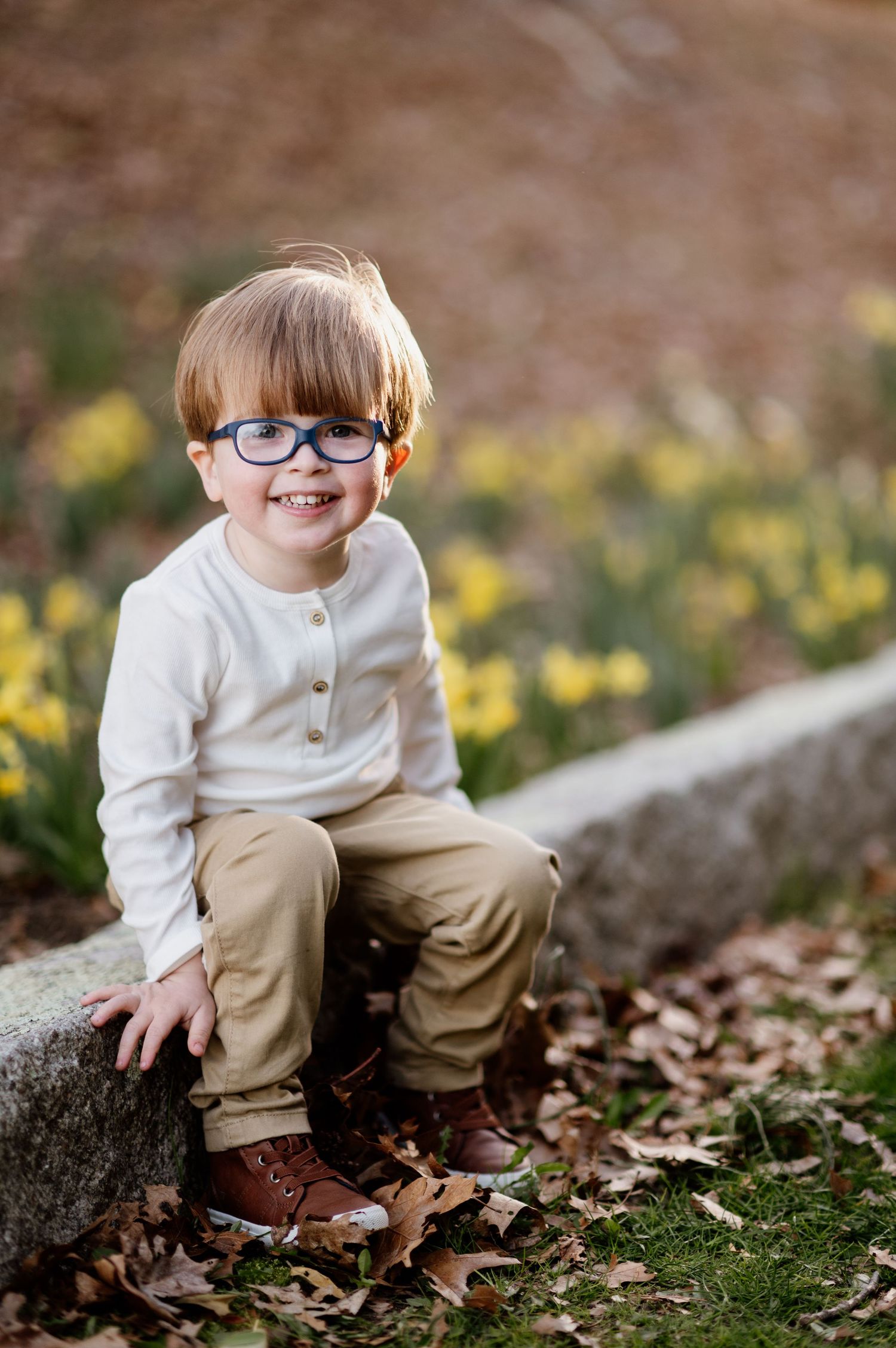 Small child in glasses and white sweater sitting on a stone wall surrounded by autumn leaves.