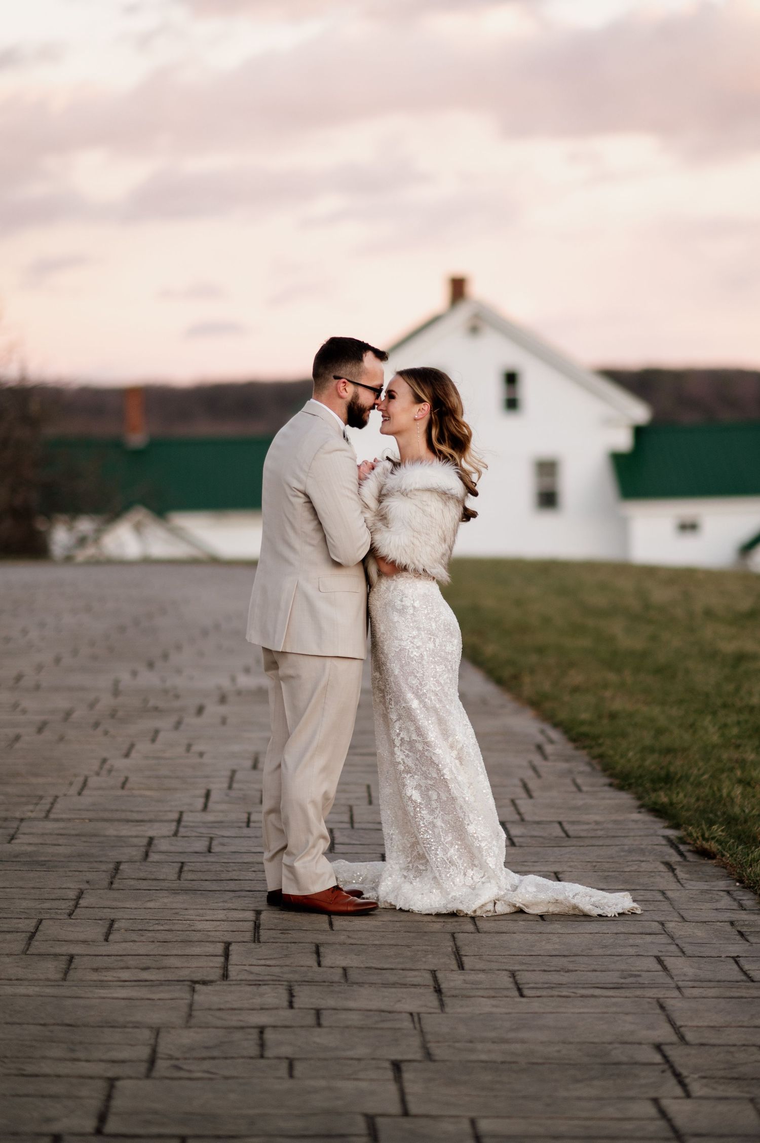 A couple shares an intimate moment on stone pavement at sunset with a farmhouse in the background.