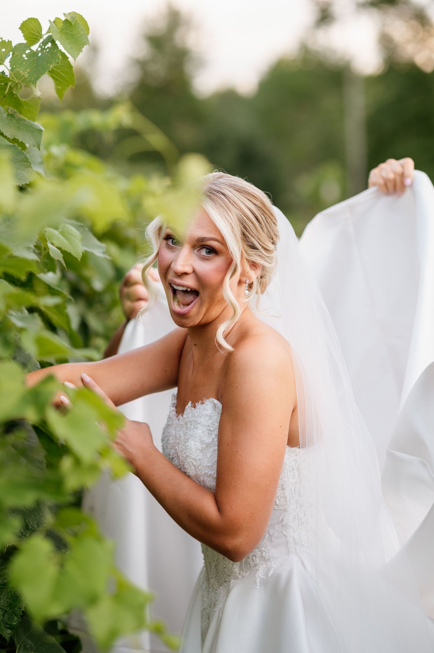 Joyful bride in white gown and veil posing playfully among lush green vineyard leaves, laughing with excitement.