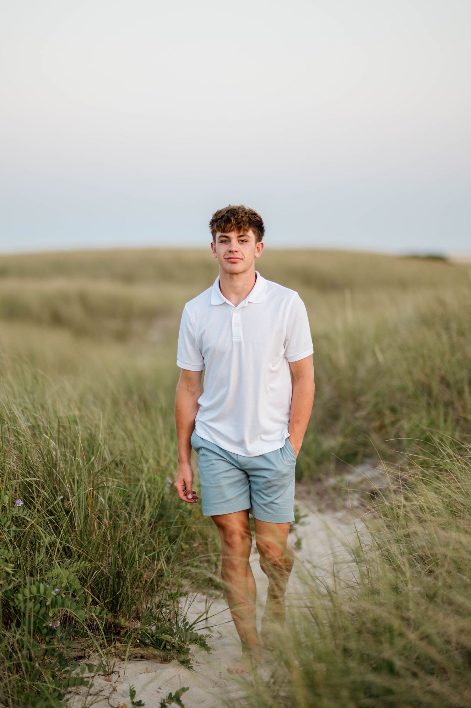 A person in a white polo shirt and light blue shorts walks through tall coastal grass on a sunny day.
