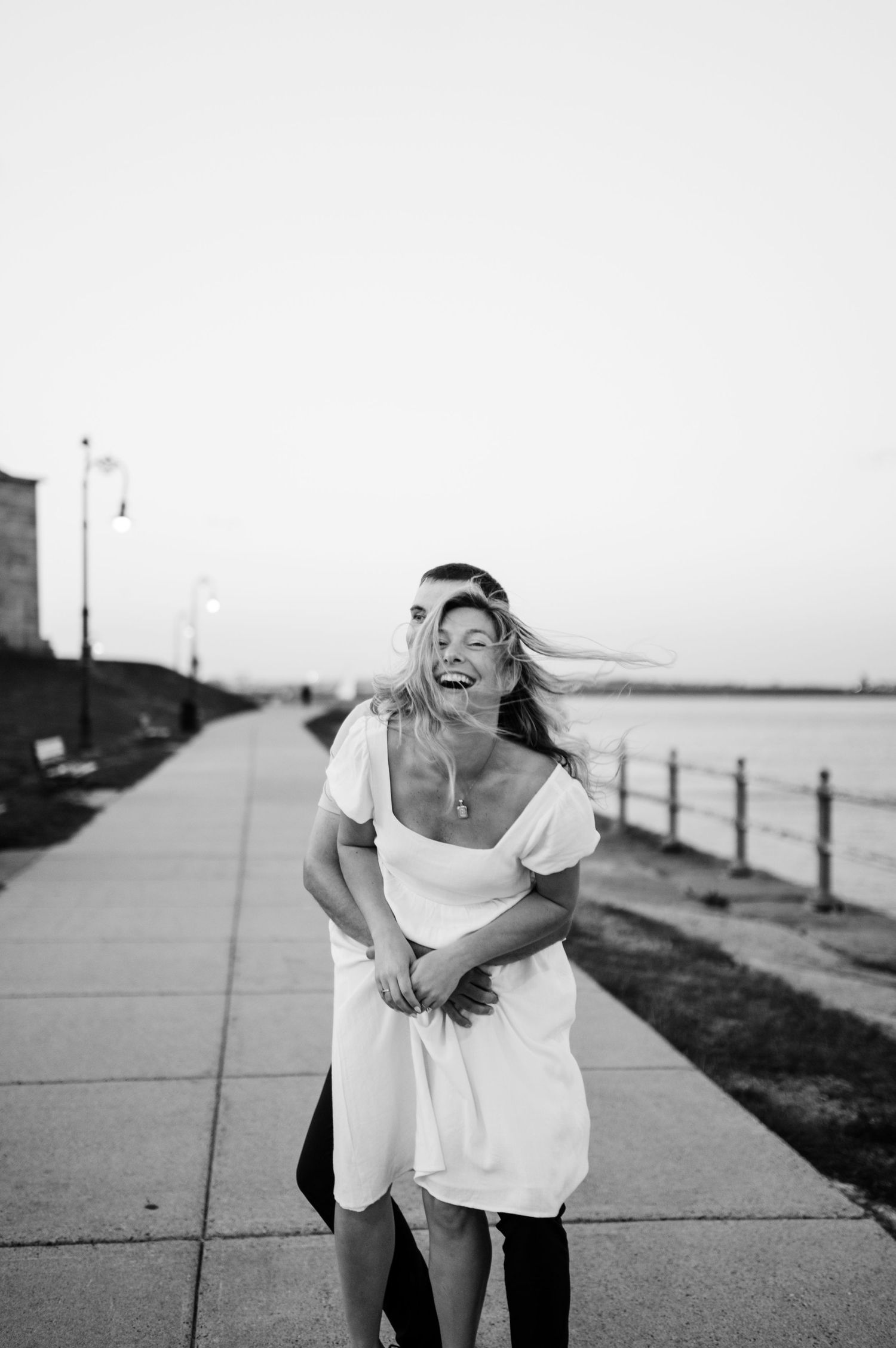 Black and white photo of a couple sharing a joyful moment on a seaside walkway.