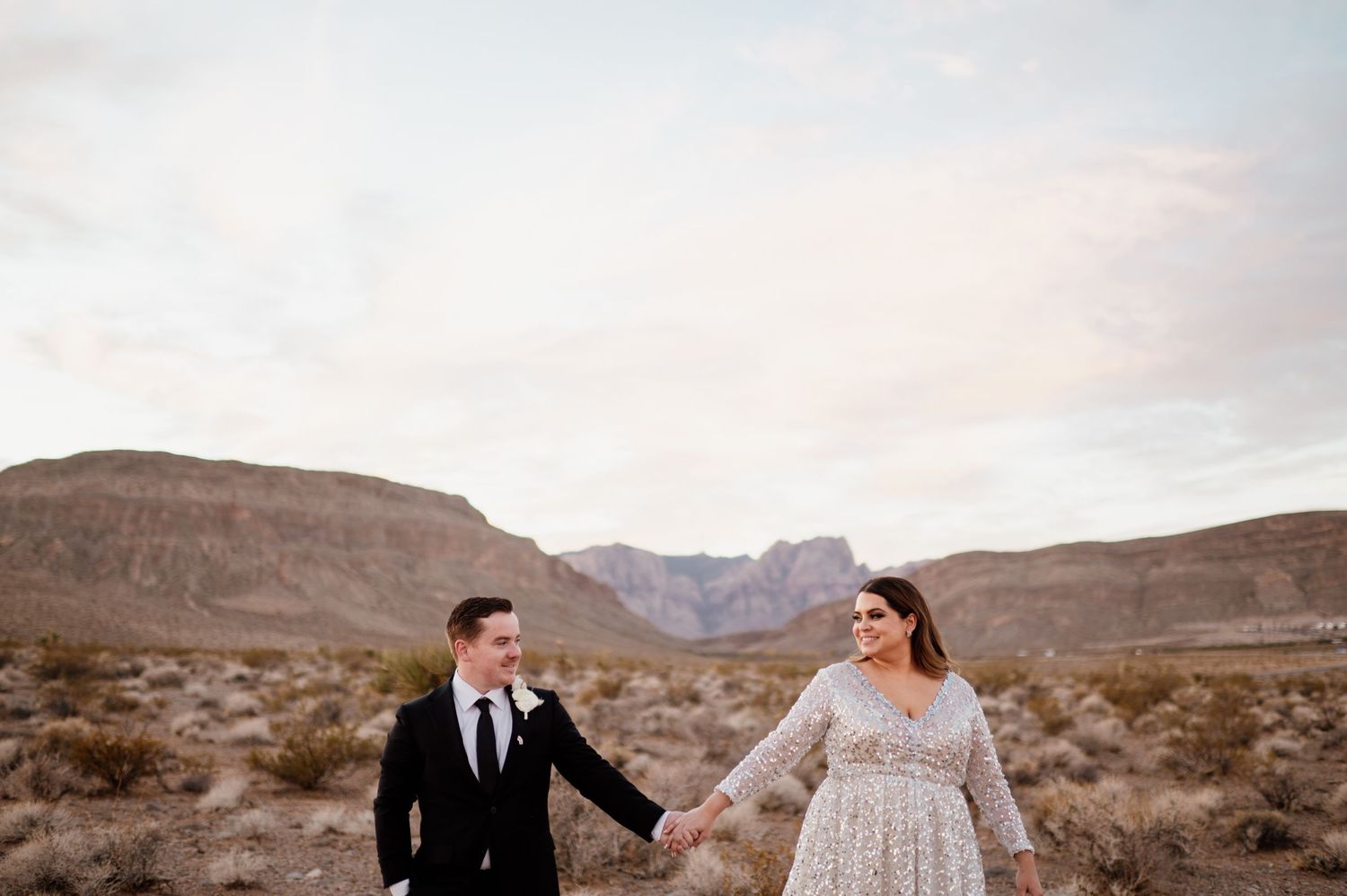 Couple in formal attire walking through a scenic desert landscape with mountains in the background at sunset.