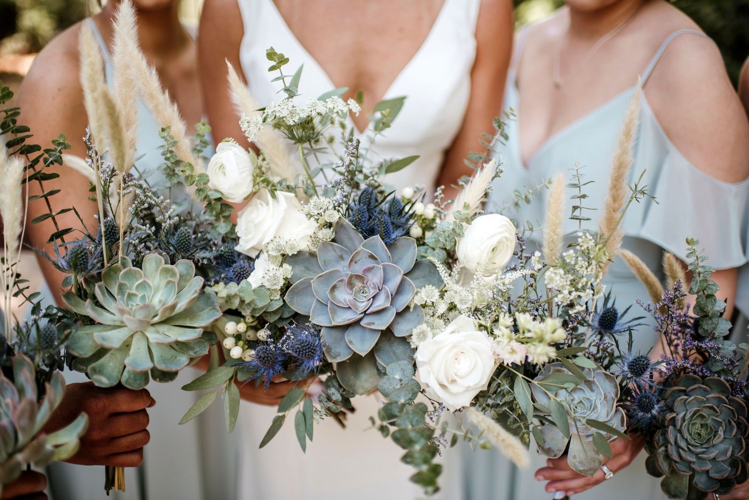 Wedding party holding elegant bouquets featuring white roses, dusty blue flowers, and pale green succulents with eucalyptus.