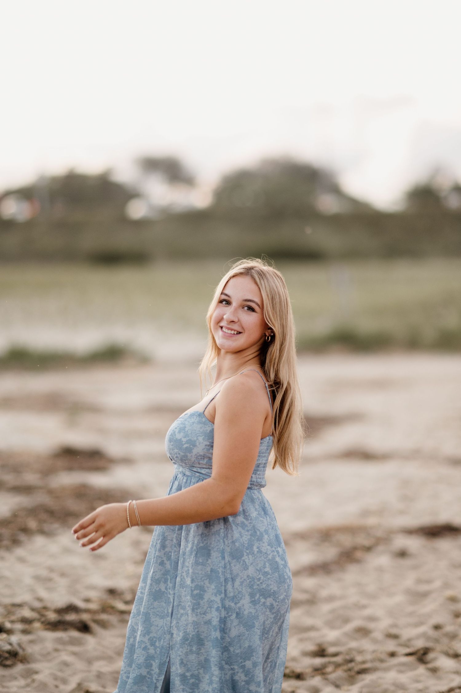 Someone in a light blue floral dress poses on a sandy beach at sunset with a natural blurred background.