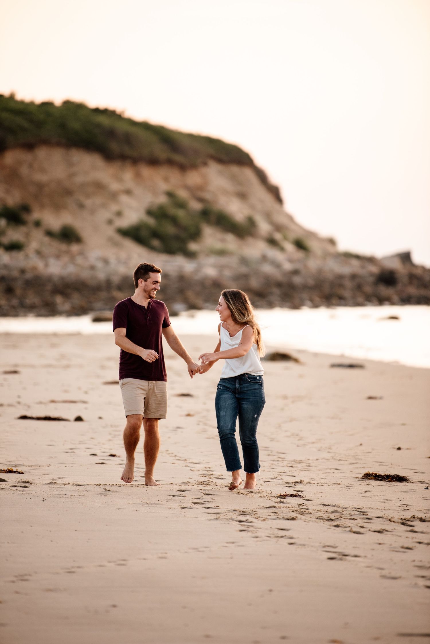 A couple walks together holding hands on a sandy beach with cliffs in the background during sunset.