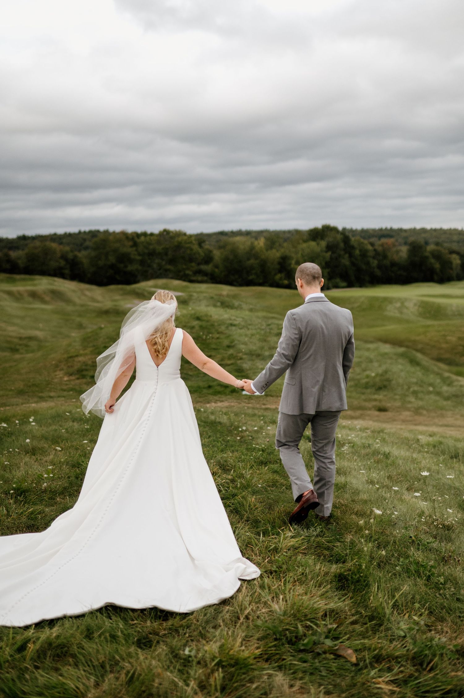 Bride's dress trails beautifully across a hilltop meadow as couple walks together.