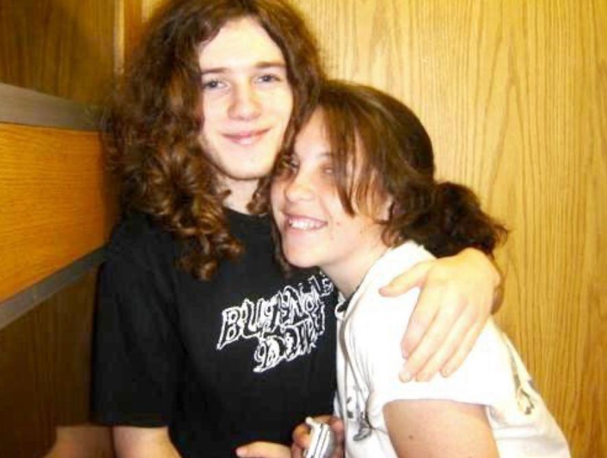 Two people with long dark hair embracing and smiling while sitting together against a wooden wall.